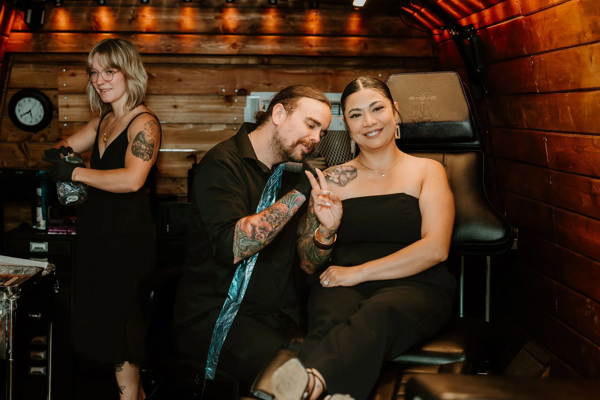 A man with tattoos and long hair is next to a woman with dark hair and a black strapless dress, seated in a chair in a wooden-paneled room. The man is smiling and making a peace sign near the woman's shoulder, who is smiling and looking at the camera