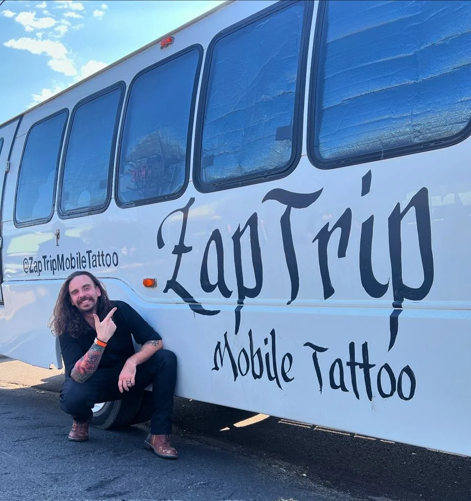 Portrait of a man with long hair and beard, squatting next to a white mobile tattoo studio bus, smiling and pointing at the studio's large sign that reads 'ZapTrux Mobile Tattoo'.