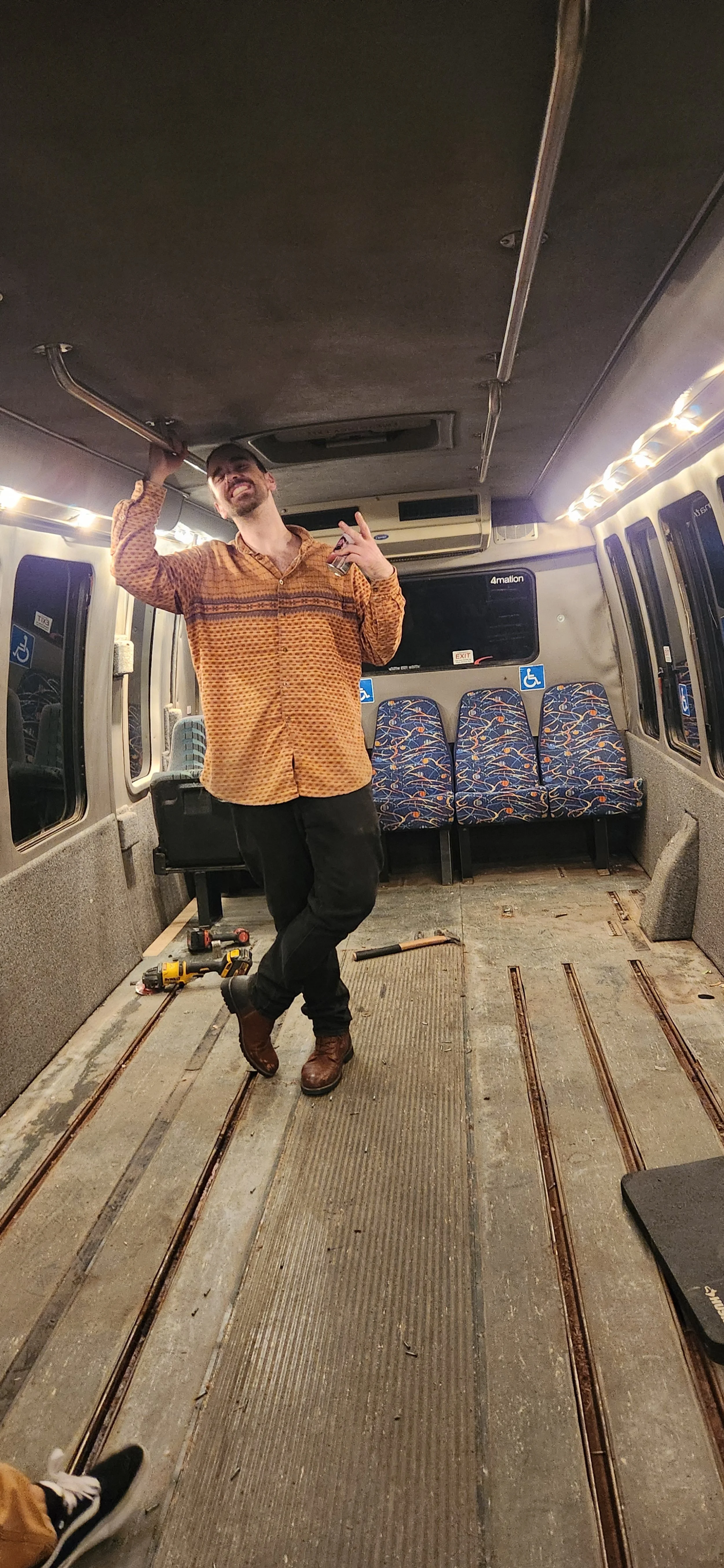 A man standing inside a partially renovated bus, smiling and gesturing happily. The bus has SCOTCH track flooring, patterned seats, and tools on the floor.