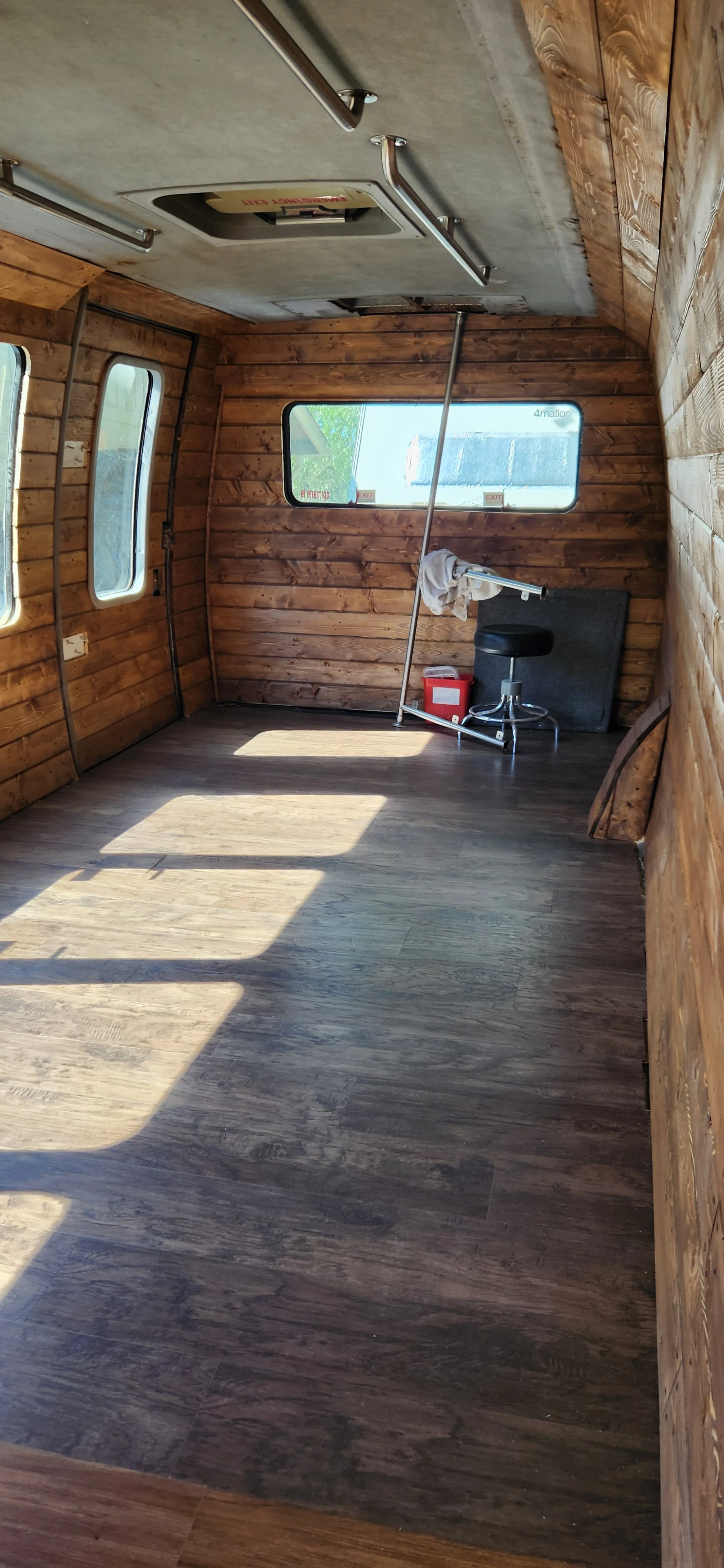 An empty room with wooden walls and floor, with sunlight casting shadows through rectangular windows, and a black stool and cleaning supplies in the corner.