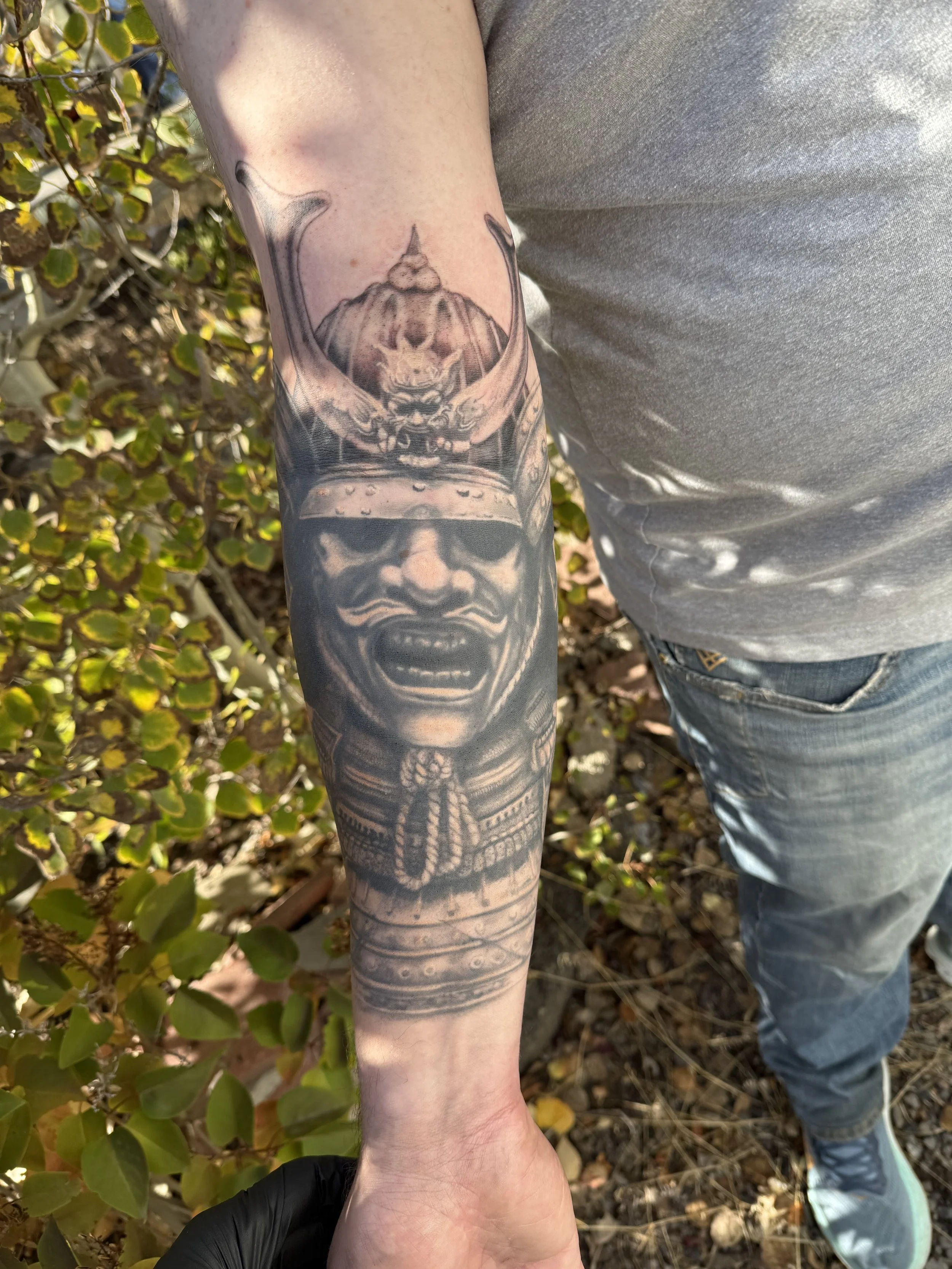 A detailed black and gray tattoo on a person's forearm featuring a fierce face in the foreground and a mythical warrior face with a horned helmet above it.