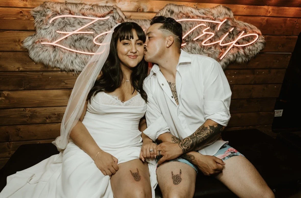 A woman in a wedding dress sits next to a man in a white shirt and shorts, both with tattoos, holding hands, with the man kissing the woman's temple. They are sitting on a black surface in front of a wooden wall with a neon sign that says 'Love Deepl