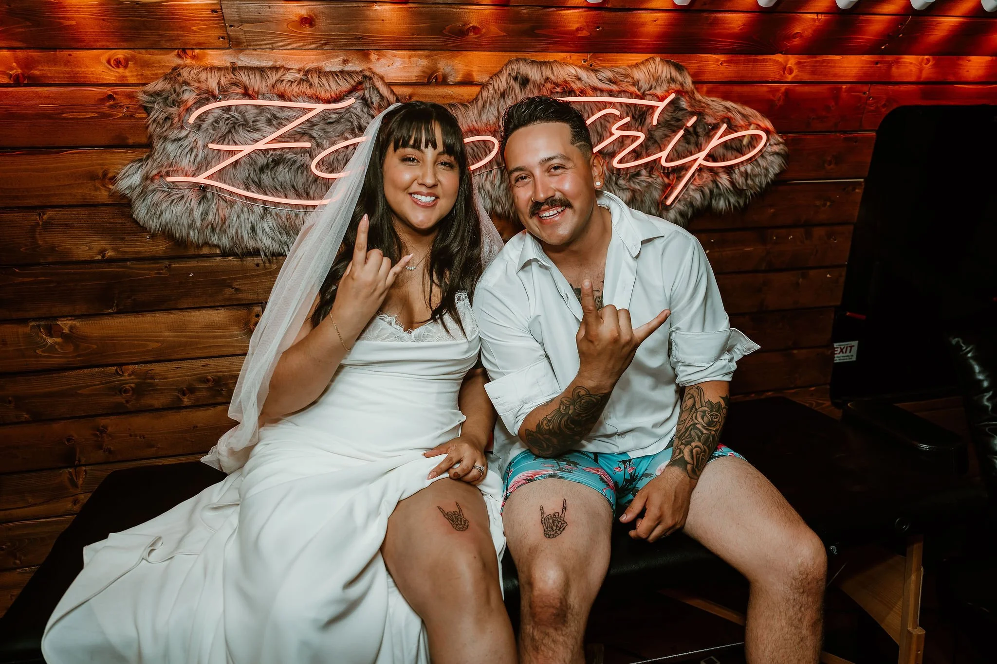 A bride and groom sitting on a bench, making hand gestures, with a wooden wall and pink neon sign that says "Zoom Up" behind them.