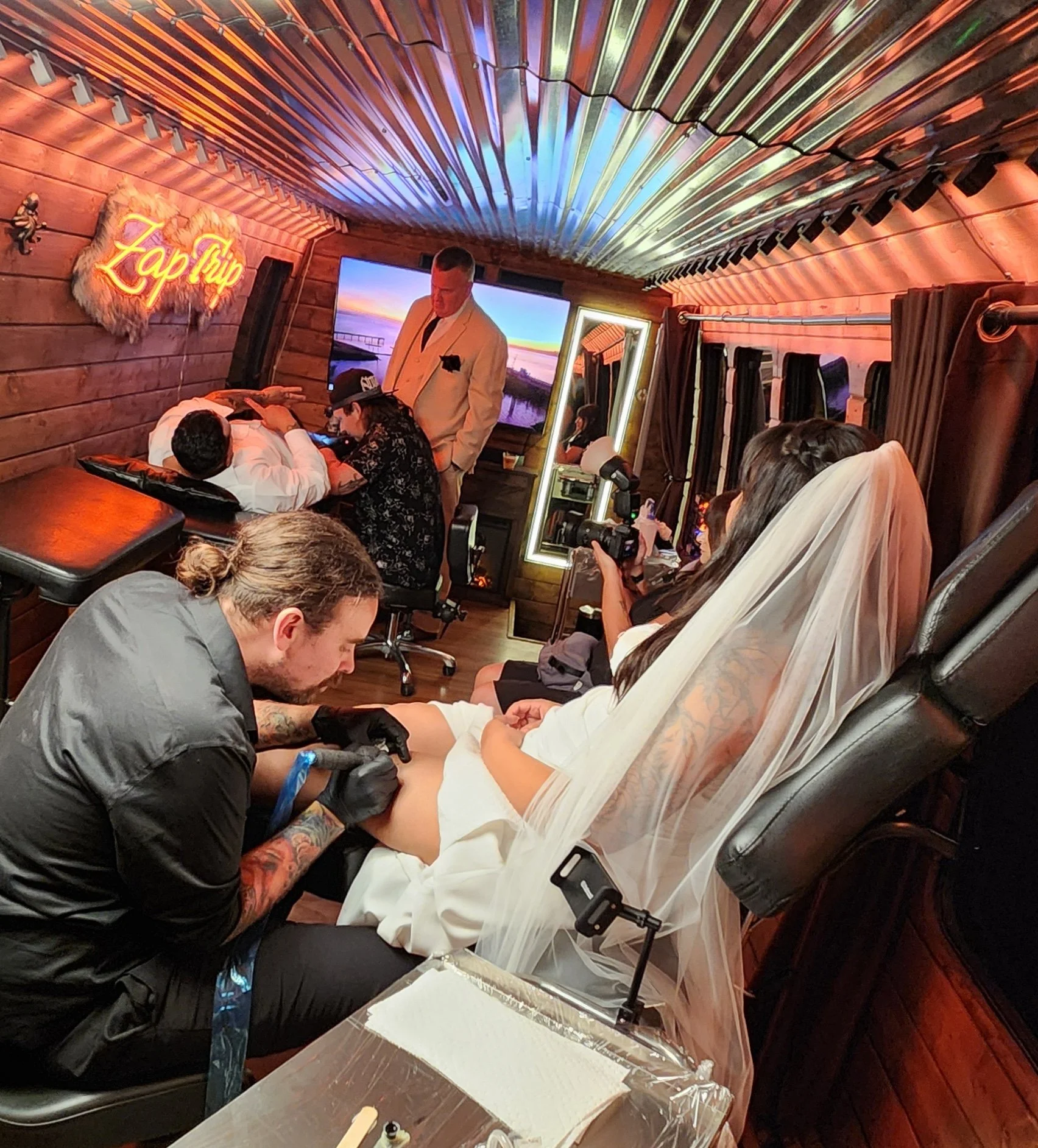 Tattoo artist tattooing a pregnant woman in a tattoo studio decorated with neon signs and sunset scenery images.