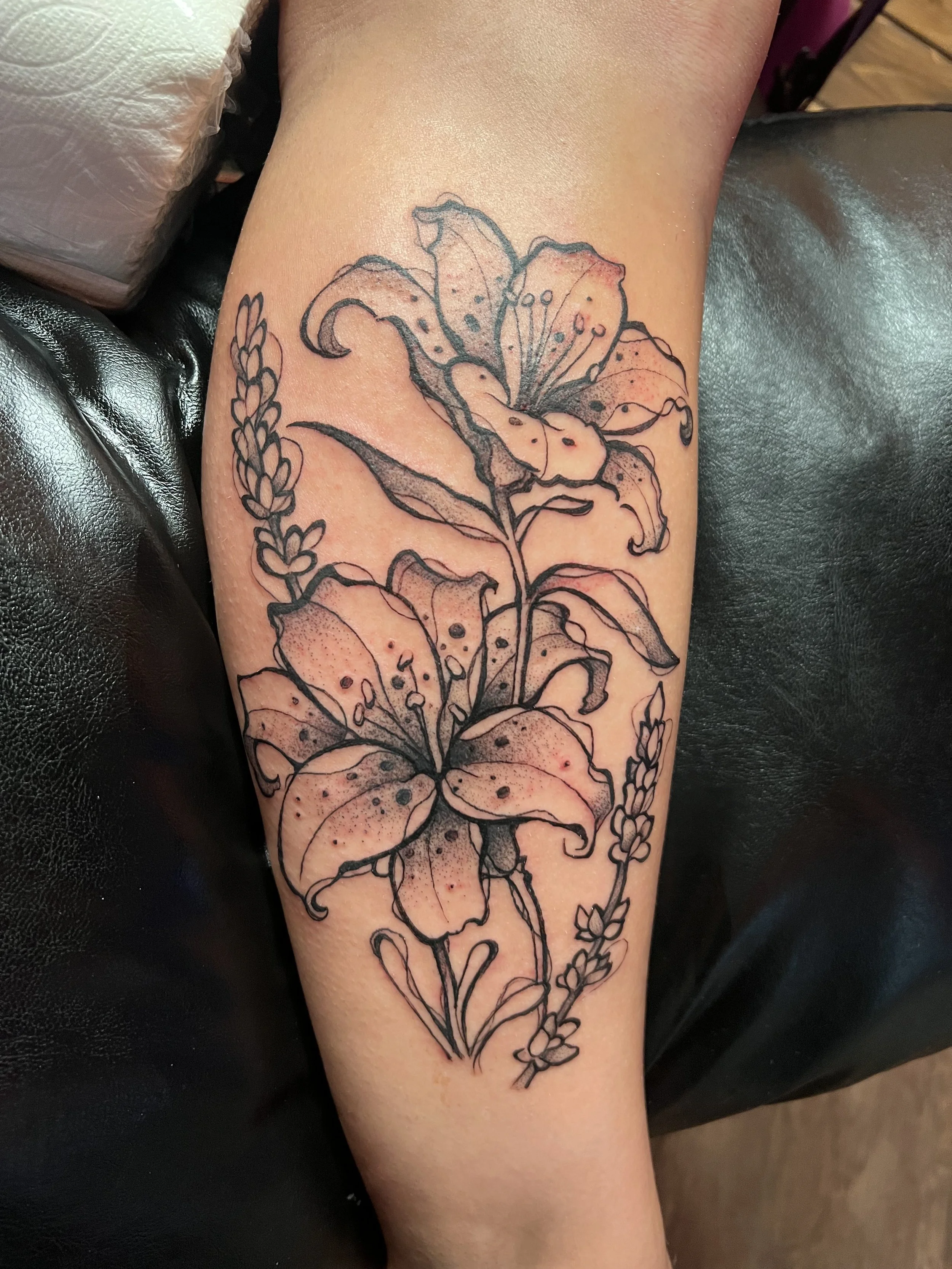 Black outline tattoo of two blooming lilies and sprigs of lavender on a person's arm.