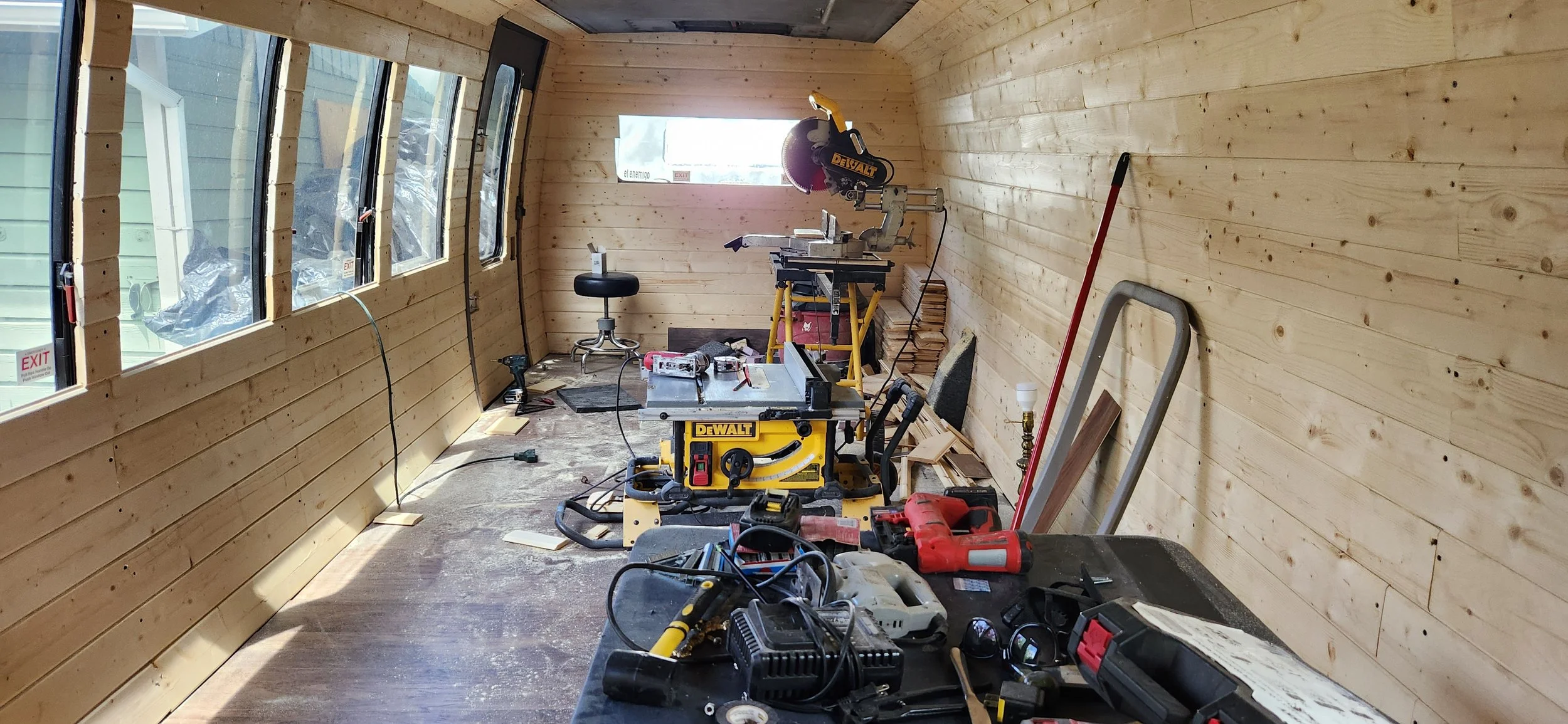 Interior of a room under construction with wooden walls and floor, featuring tools and equipment such as saws, drills, and a ladder, and large windows along one side.