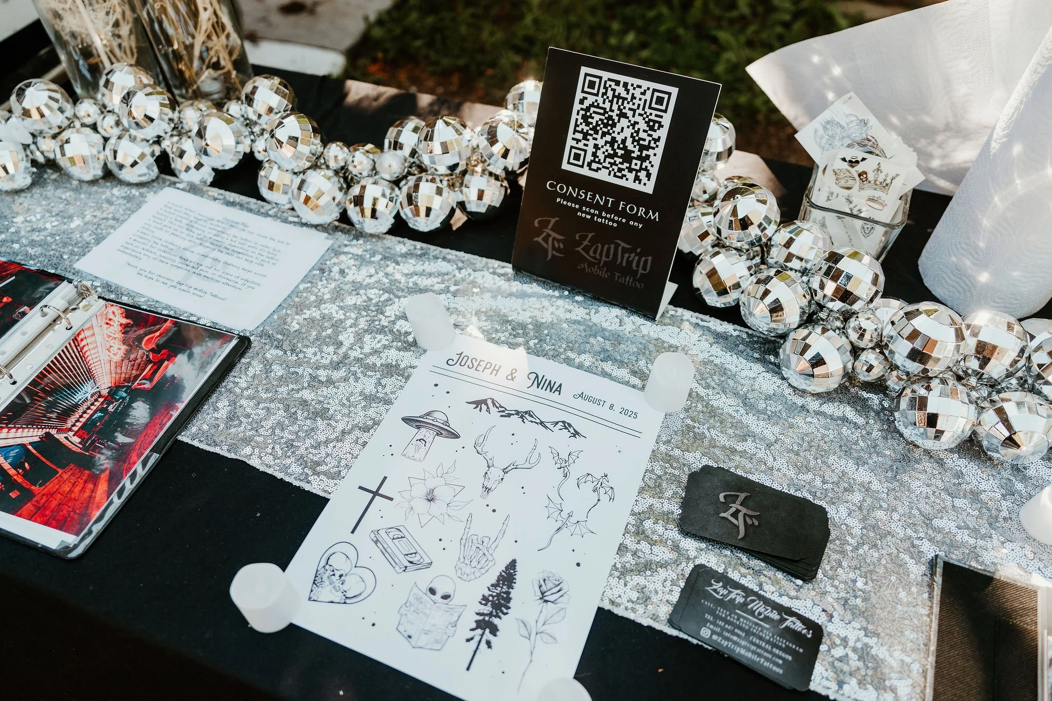 A table decorated with a silver sequin table runner, featuring tattoo designs, a consent form with a QR code, business cards for ZapTrip Tattoo, silver disco ball garland, a pamphlet or flyer, and various tattoo related items.