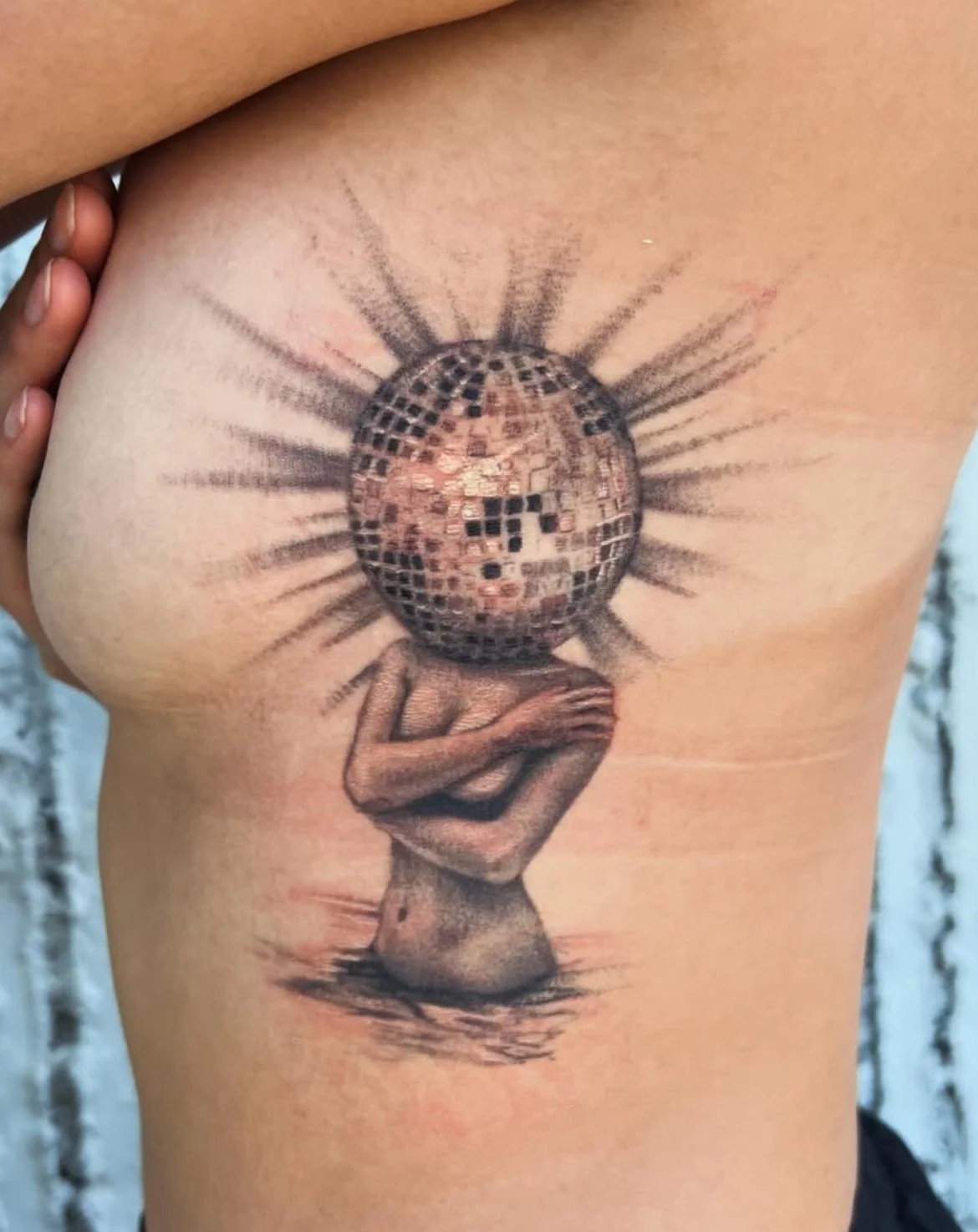 Tattoo of a person with a body holding a large disco ball-headed sphere with rays emitting from it, covering the face, on a person's side ribcage.