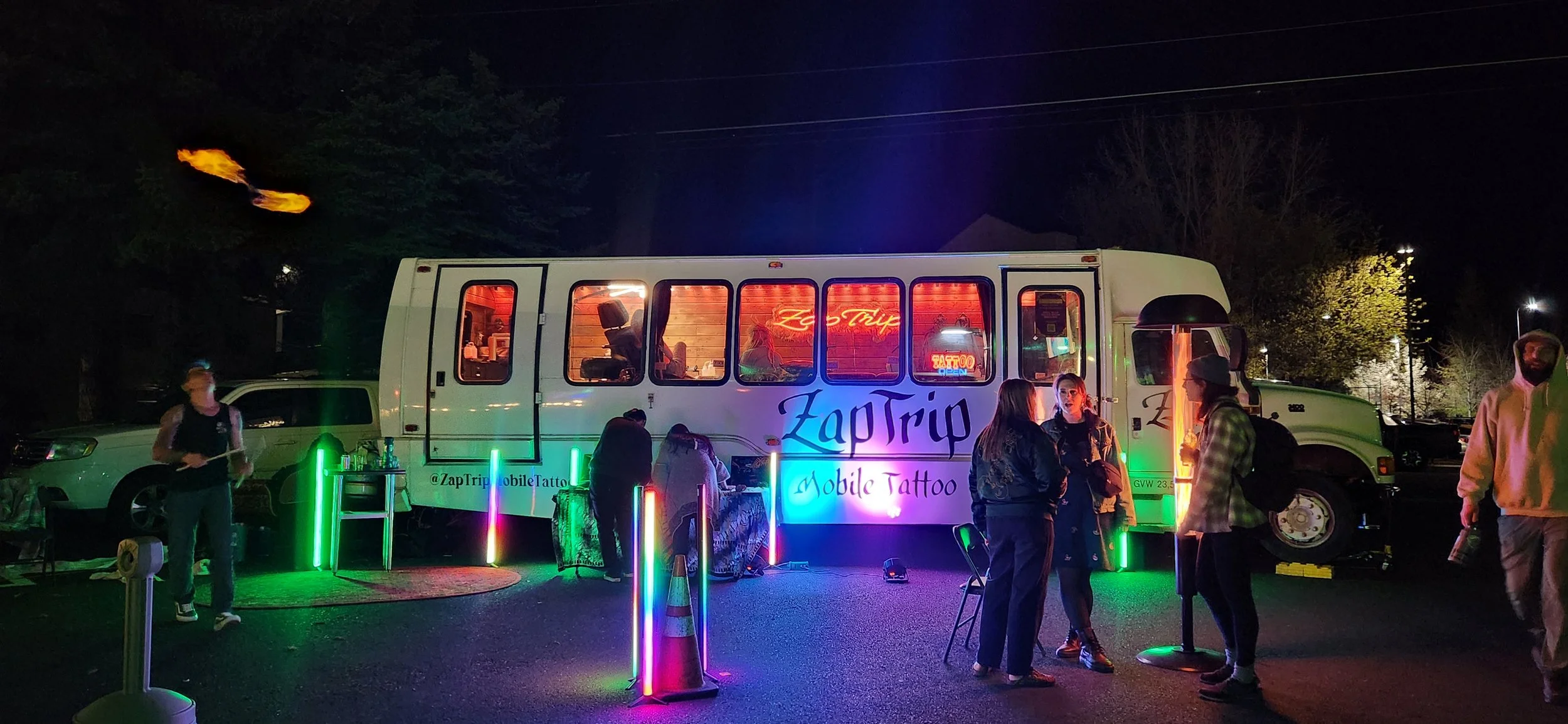 A mobile tattoo parlor called "ZapTrip" is set up outdoors at night with neon lights and several people gathered around. The tattoo truck has a large window with a warm inside glow and is decorated with colorful vertical light columns, chairs, and ta