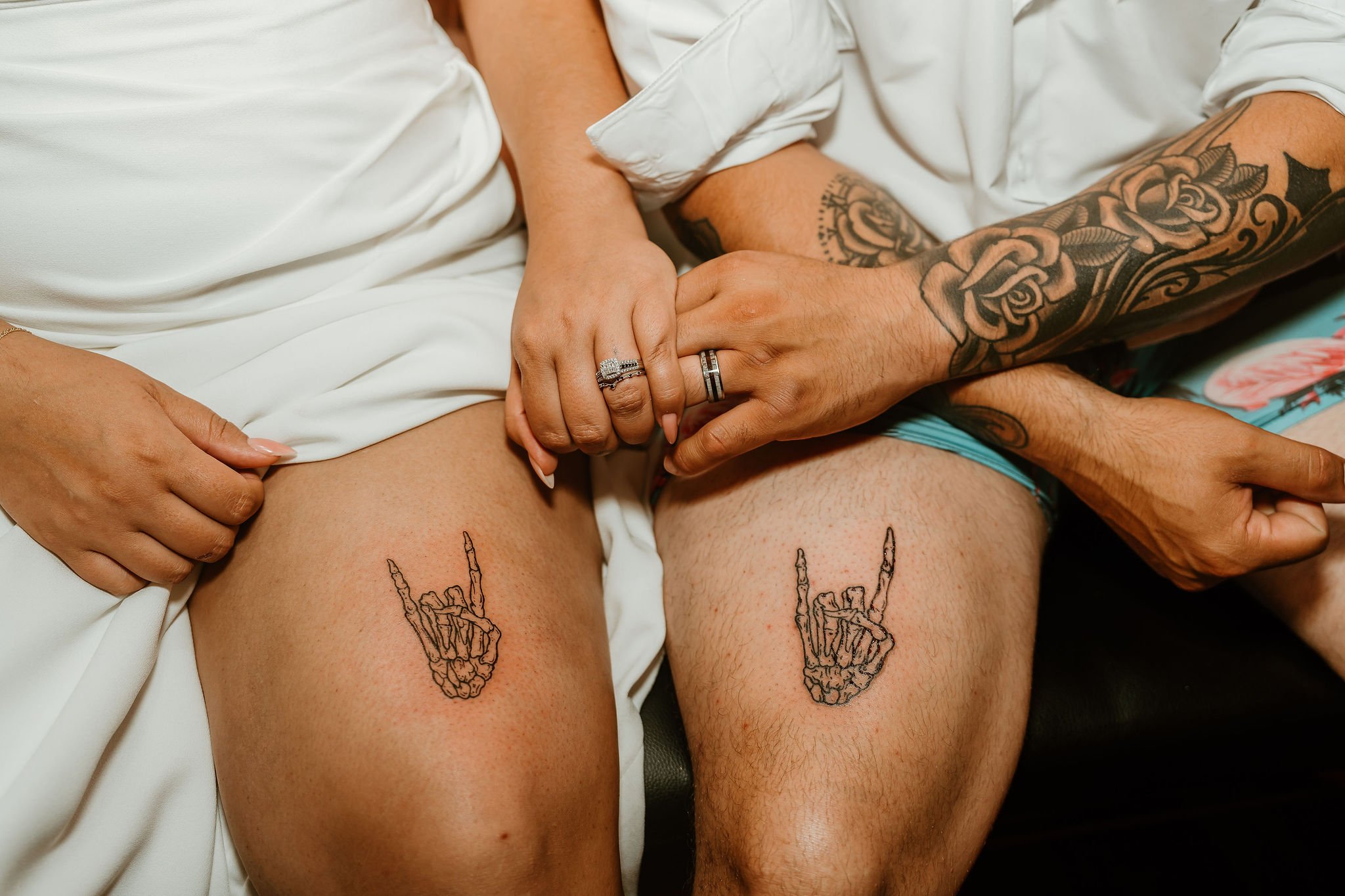 A couple holding hands and displaying matching tattoos of a skeleton hand making the 'rock on' gesture on their thighs, with the woman's hand wearing rings.