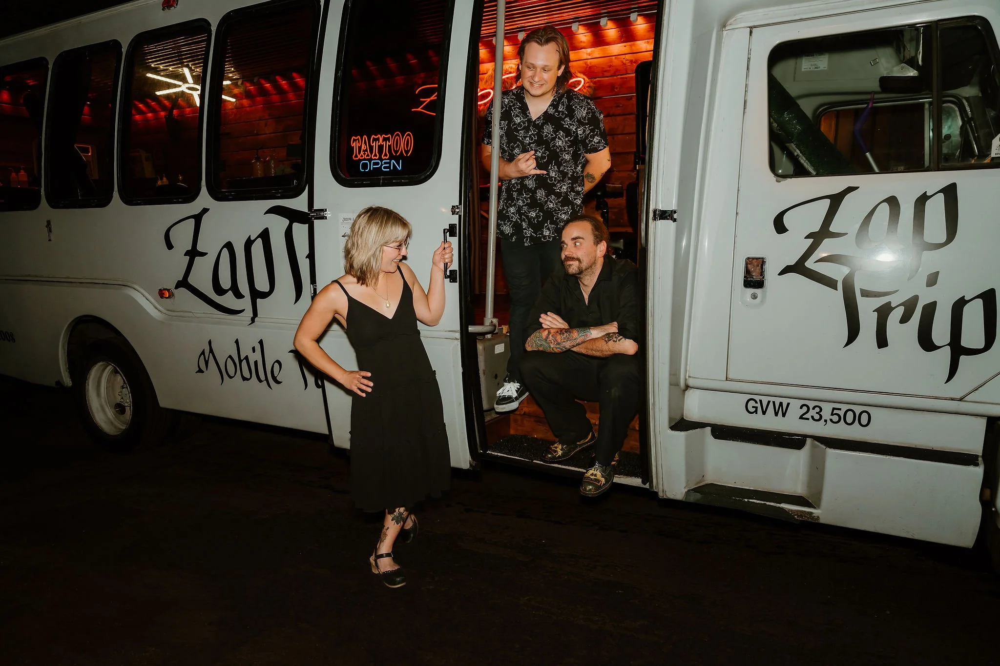 Three people around a mobile tattoo bus called Zap Tripp, with a neon tattoo sign illuminated in the background. One person is sitting on the bus steps, another stands outside, and the third is standing on the bus steps, looking at their phone.