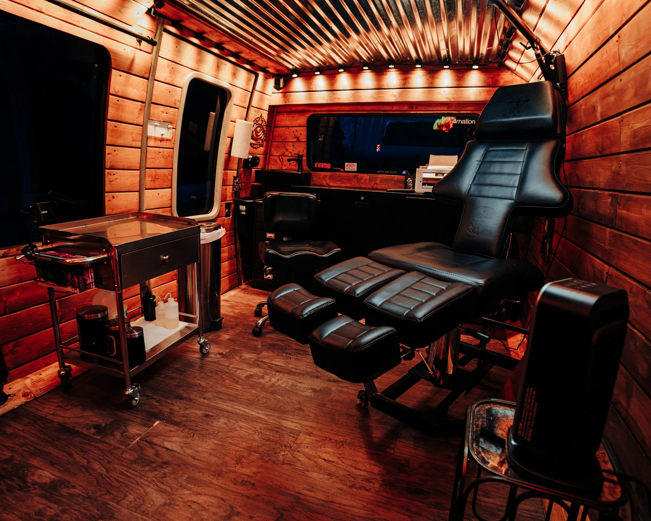 Inside a wooden-paneled room with a black leather reclining chair, a small rolling cart with medical supplies, and a wood floor, potentially a mobile medical or tattoo parlor space.