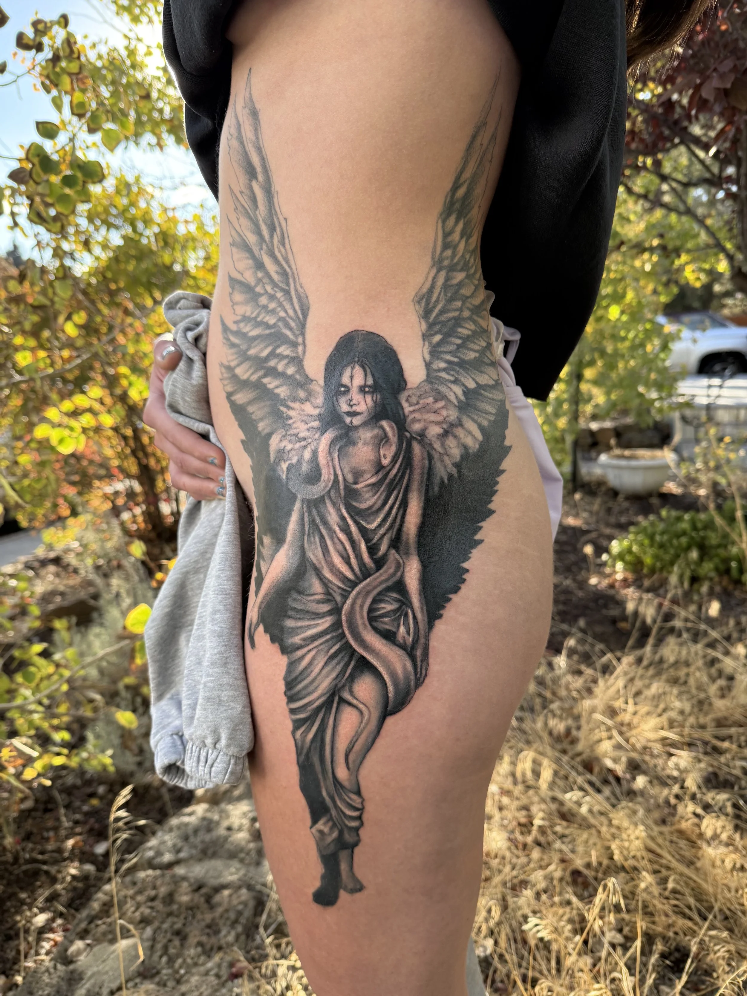 A tattoo of a mythological or fantasy creature on the person's thigh, featuring a woman with dark hair, angelic wings, and a snake wrapped around her leg, in a grayscale style.