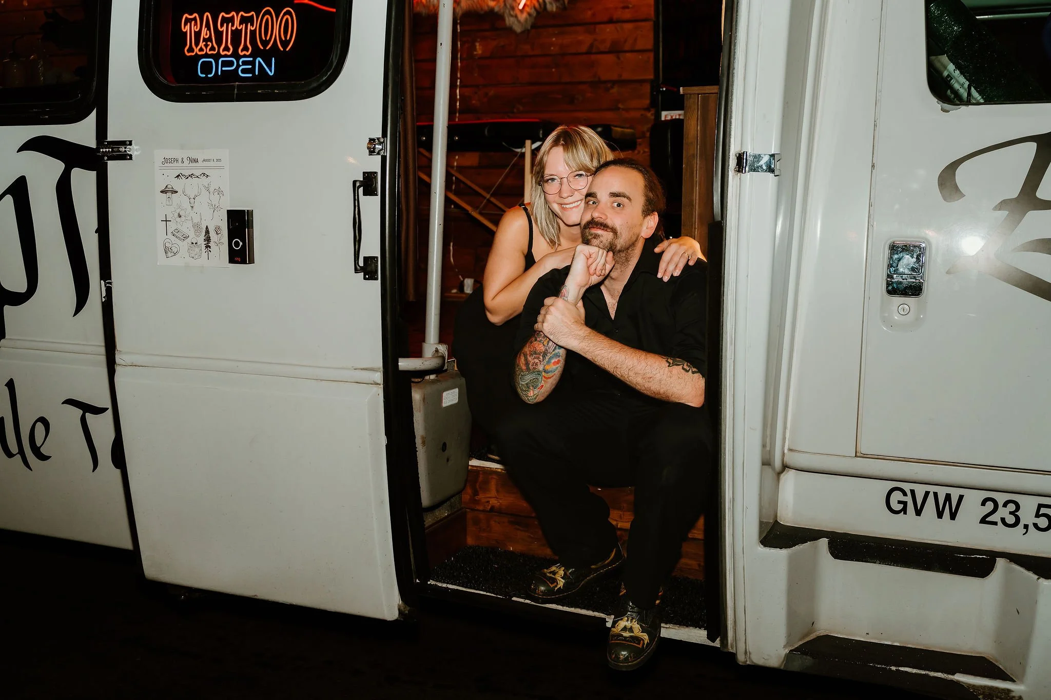 A woman and a man sitting in the doorway of a tattoo truck, smiling. The woman has blonde hair and glasses, and the man has tattoos on his arm, dark hair, and a beard.