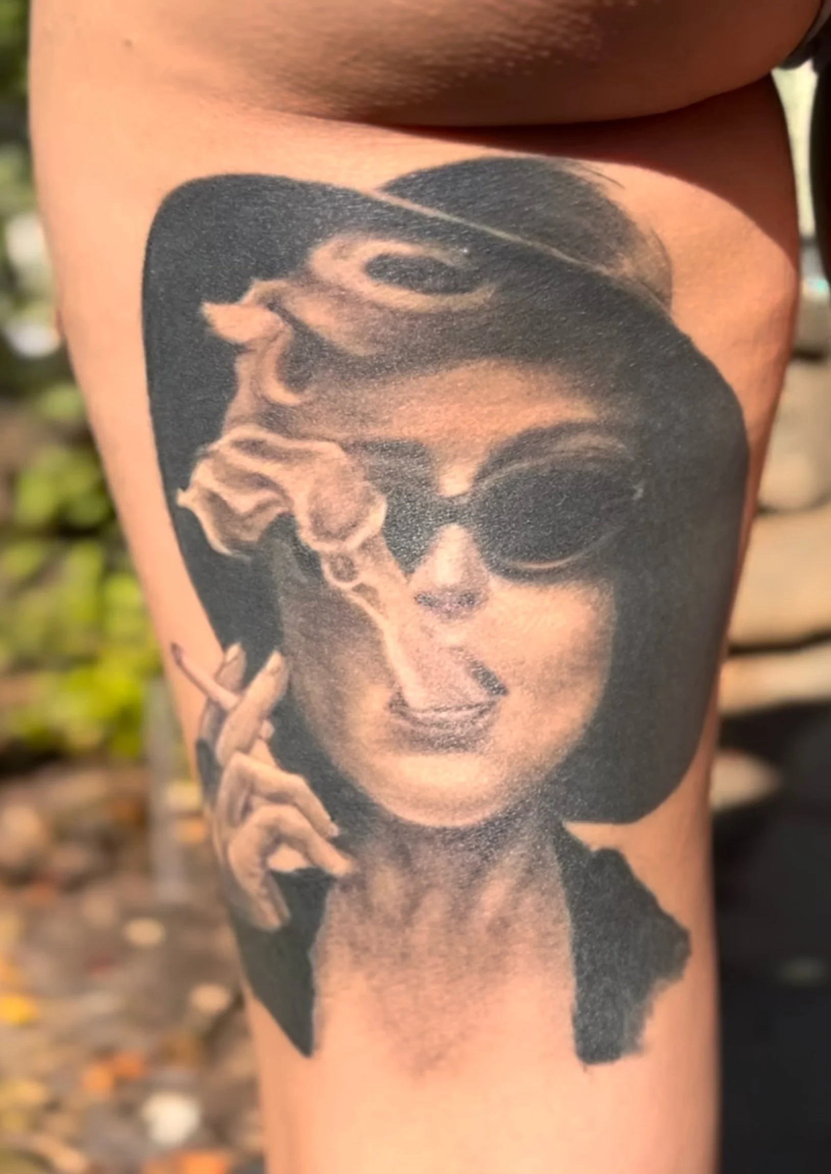 A black and gray tattoo of a woman wearing sunglasses, holding a cigarette with smoke rising, and wearing a headband.