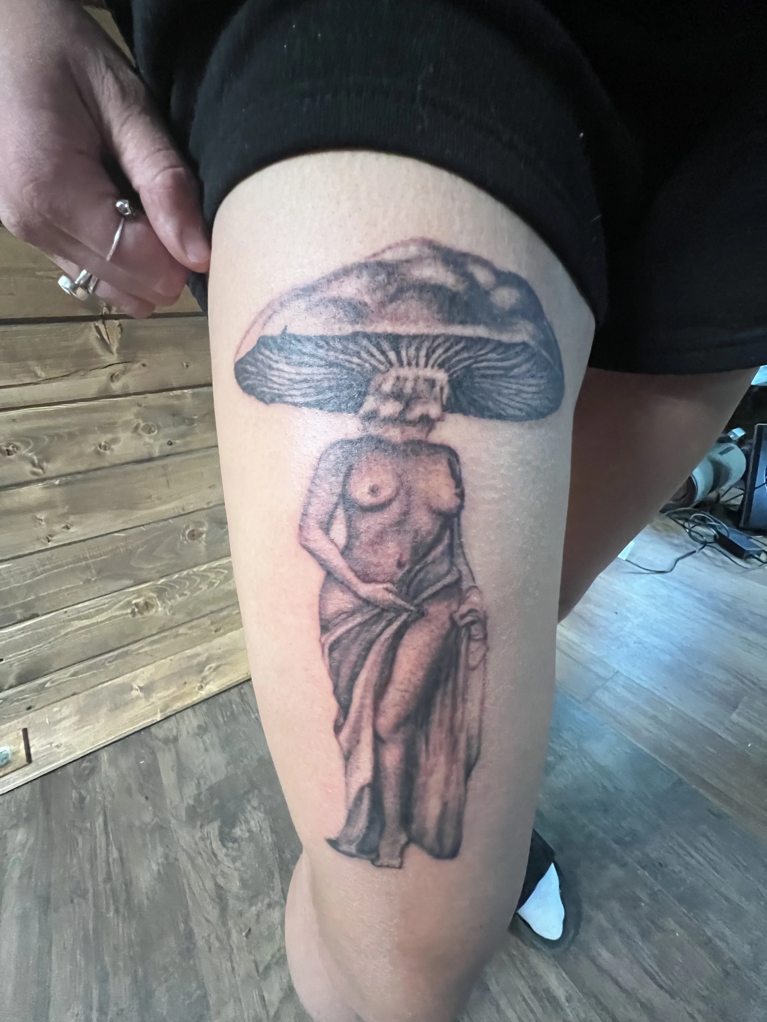 Tattoo of a woman with a large mushroom cap as her head, standing with a cloth around her waist, on a person's thigh.