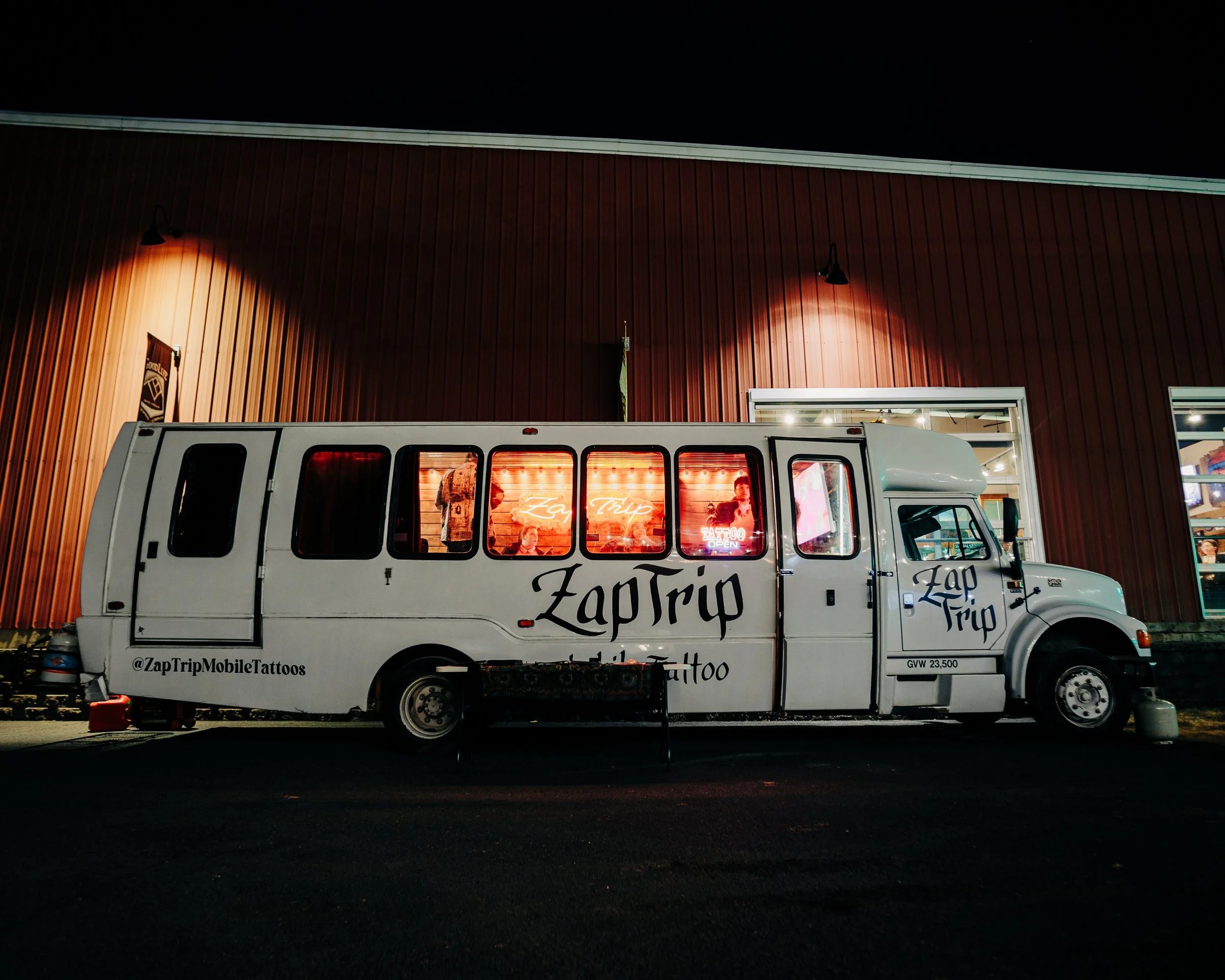 A white vehicle with the words 'Zap Trip' and '@ZapTripMobileTattoos' written on the side, parked in front of a building with a wooden exterior at night. The vehicle has large windows revealing people inside and neon signs.