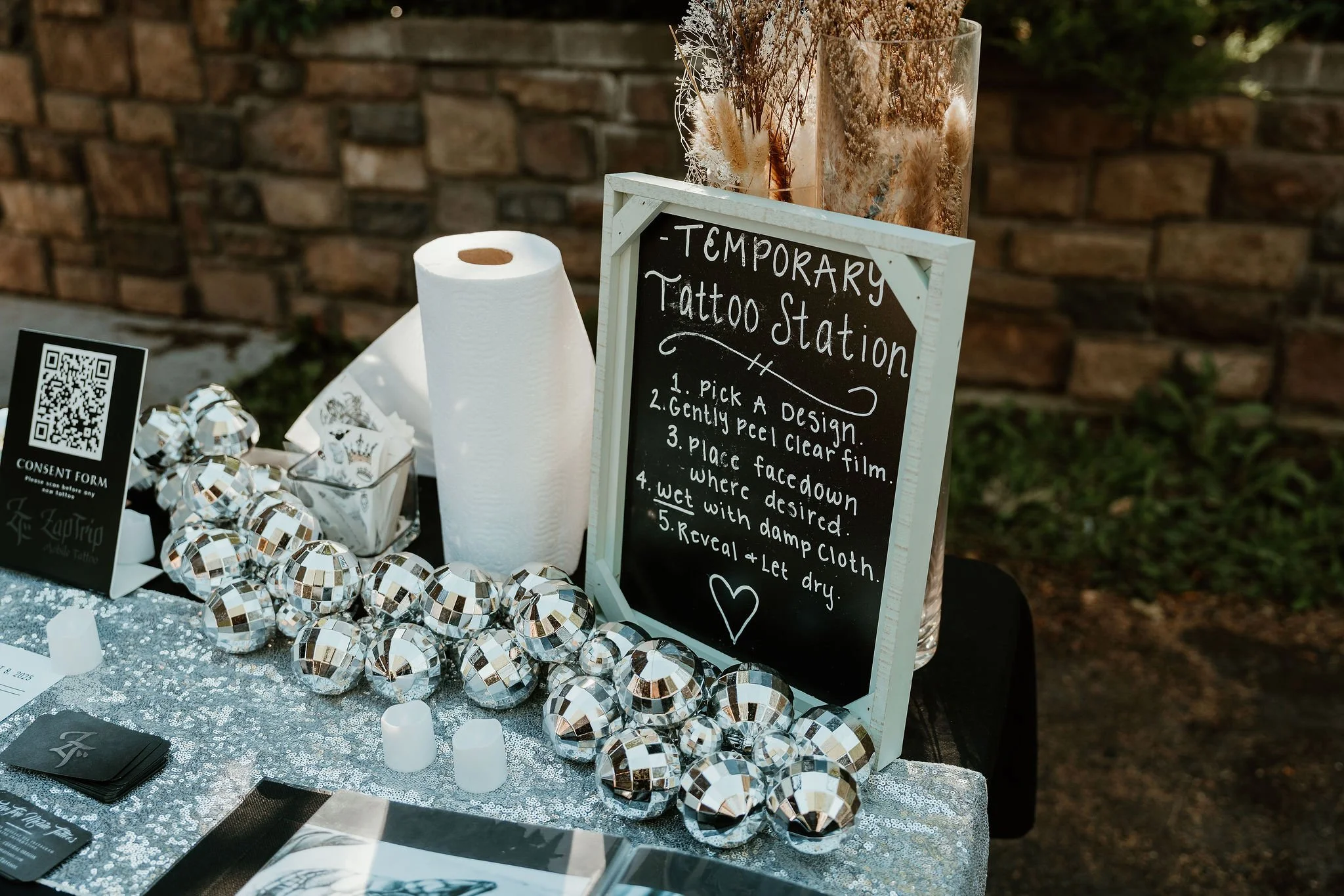 A sign at a temporary tattoo station with instructions for getting a tattoo, placed on a table decorated with shiny silver spheres, candles, and a roll of paper towels, with a brick wall and outdoor greenery in the background.