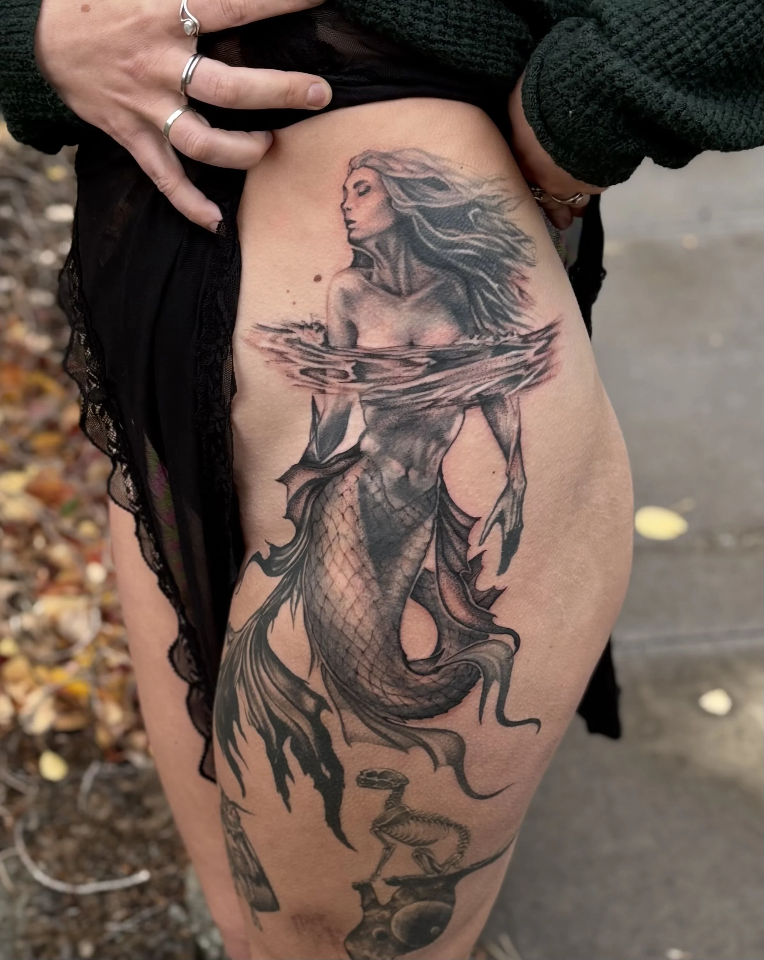 A tattoo of a mermaid with long flowing hair, partially submerged in water, with a scaled fish tail, on a person's upper thigh. There are additional decorative elements below.