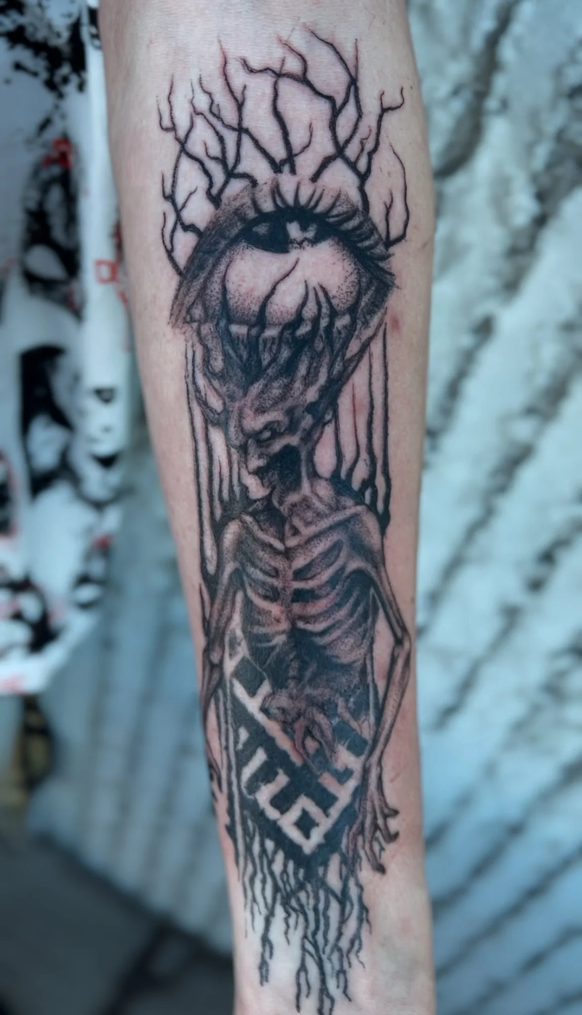 Black and gray tattoo of a skeletal figure with exposed ribs, with a large eye above it, and abstract, tree-like branches extending from the eye and head, with dripping lines and a banner with text underneath.