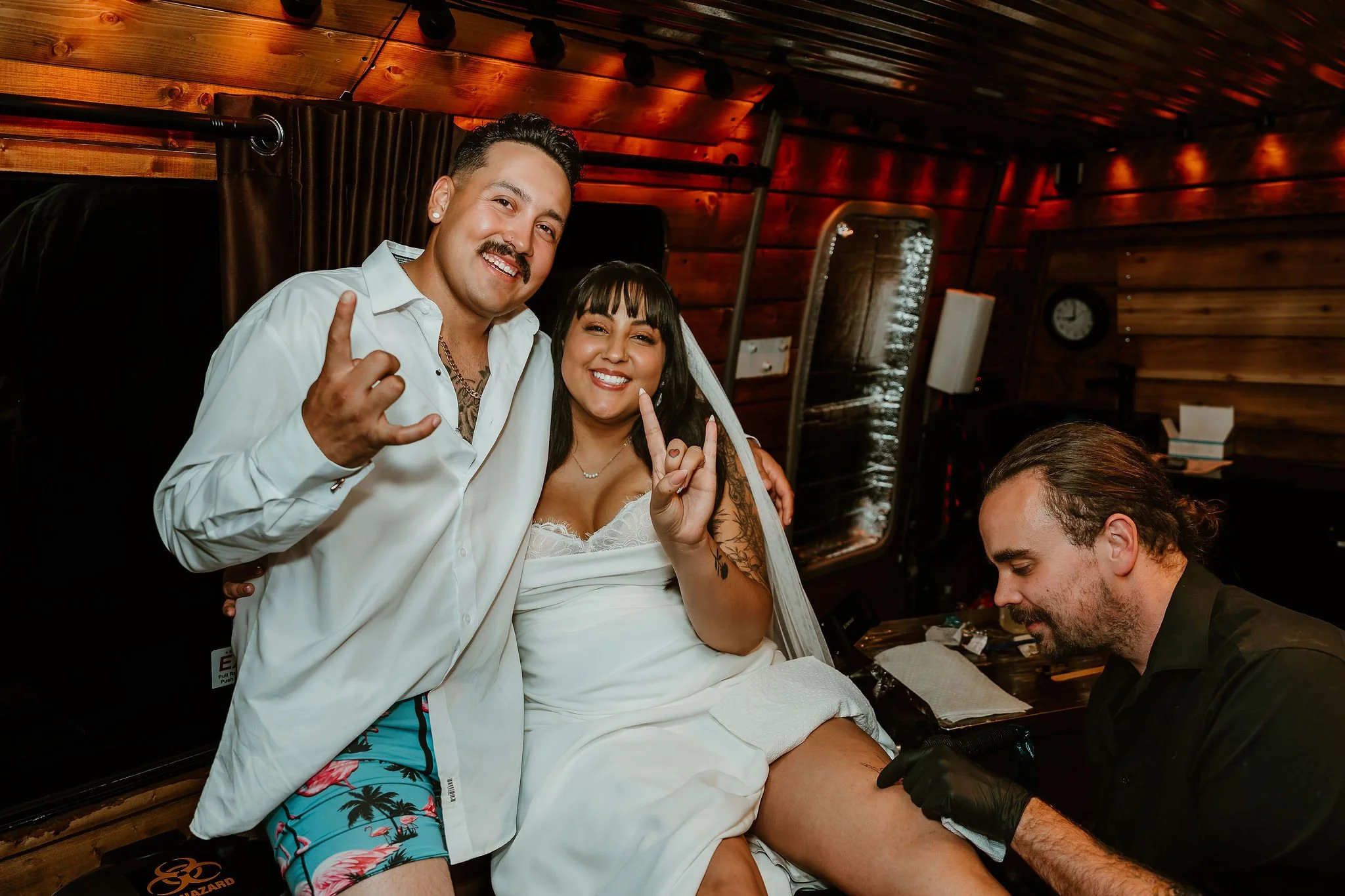 A couple, a bride and groom, smiling and making peace signs with their fingers at a tattoo station, with the groom wearing a white shirt and Hawaiian shorts, and the bride in a wedding dress. A tattoo artist is working on the bride's thigh in a rusti