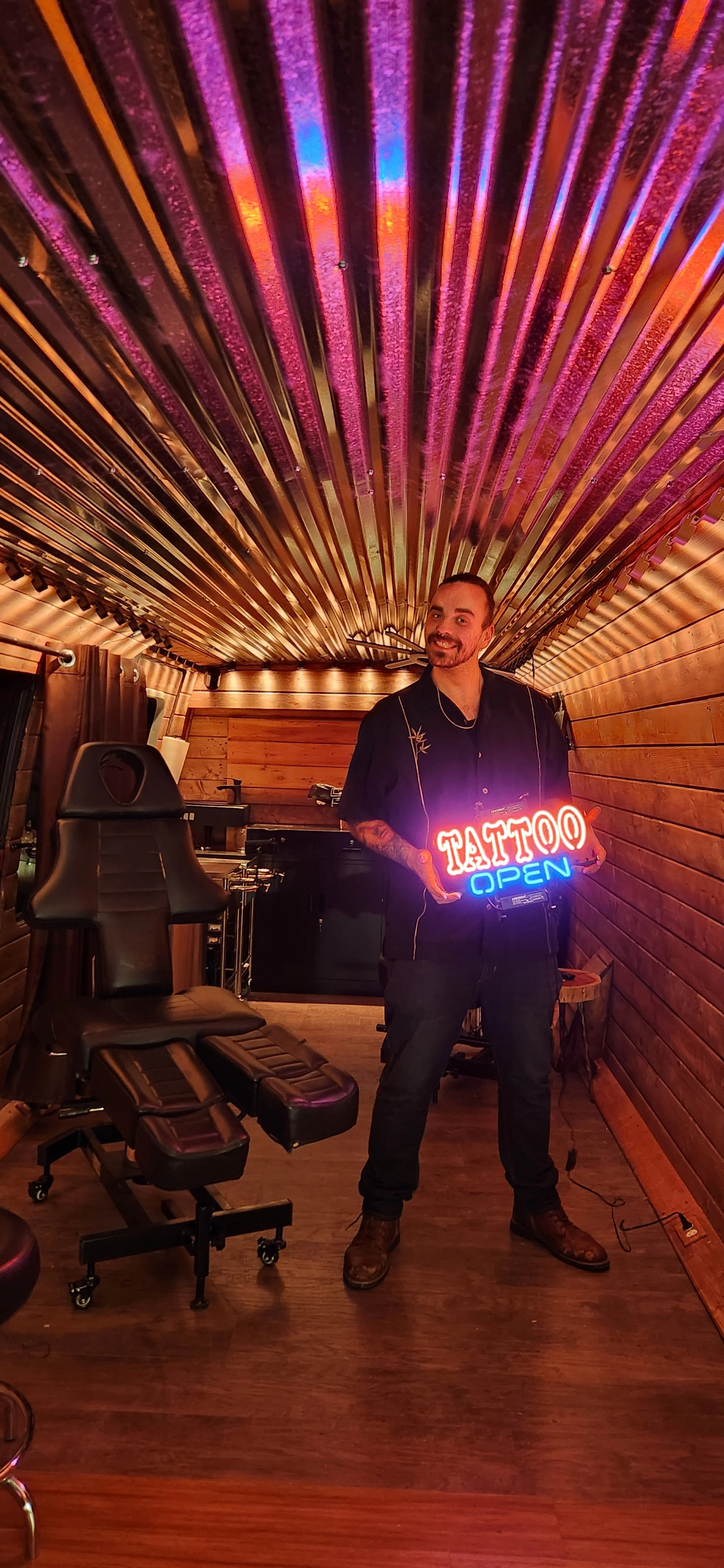A man with a beard and tattoos smiling while holding a neon sign that reads 'TATTOO OPEN' in a wooden tattoo studio.