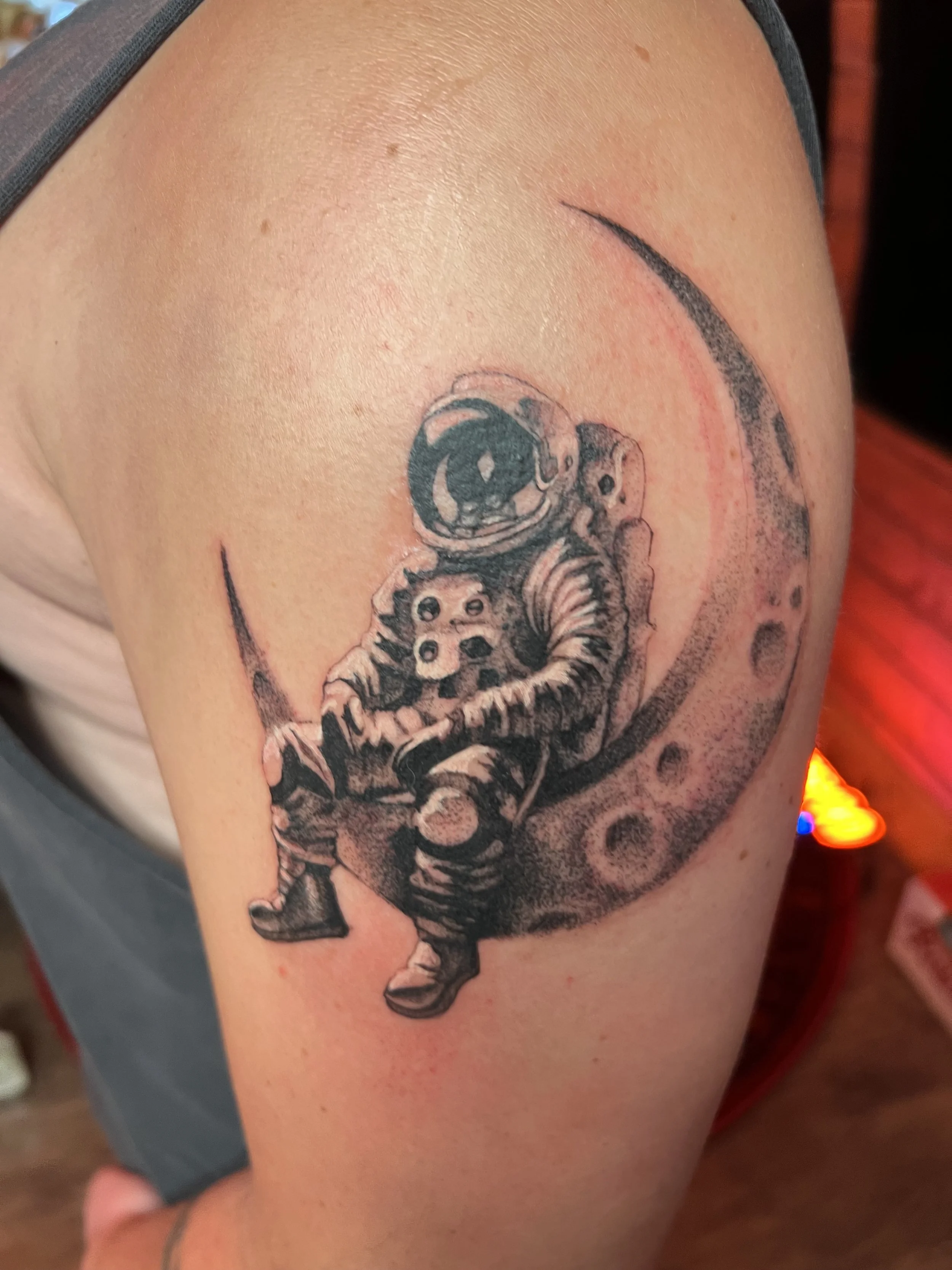 Tattoo of an astronaut sitting on the moon in black ink, with detailed shading and a crescent moon in the background.