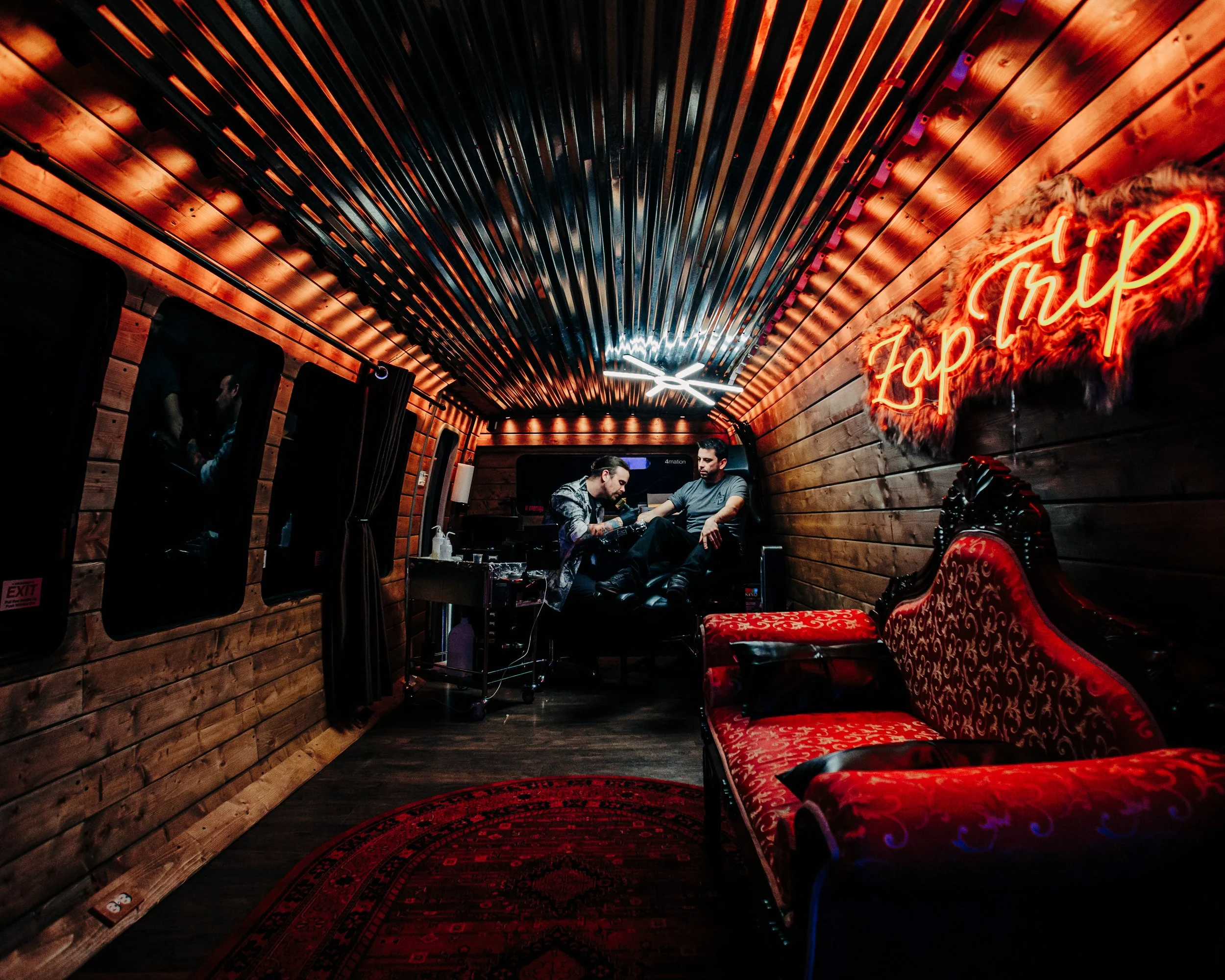 A tattoo studio interior with warm wooden walls and ceiling, a red velvet vintage sofa, a neon sign reading 'Tattoo' on the wall, and two men seated on a tattoo chair with one providing a tattoo to the other, illuminated by ambient lighting.