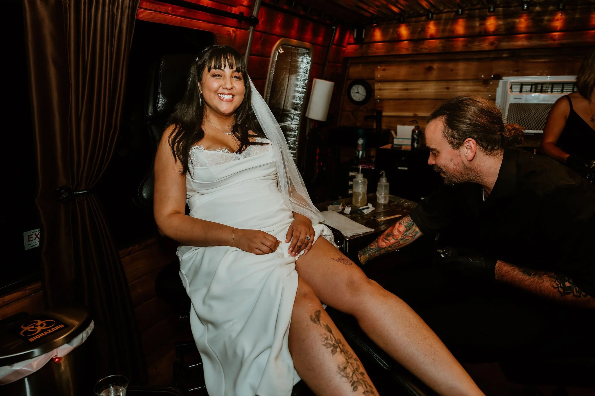 A woman in a white dress and veil smiles while sitting in a tattoo parlor chair as a man tattoos her right thigh. The room has wooden walls and dim lighting.