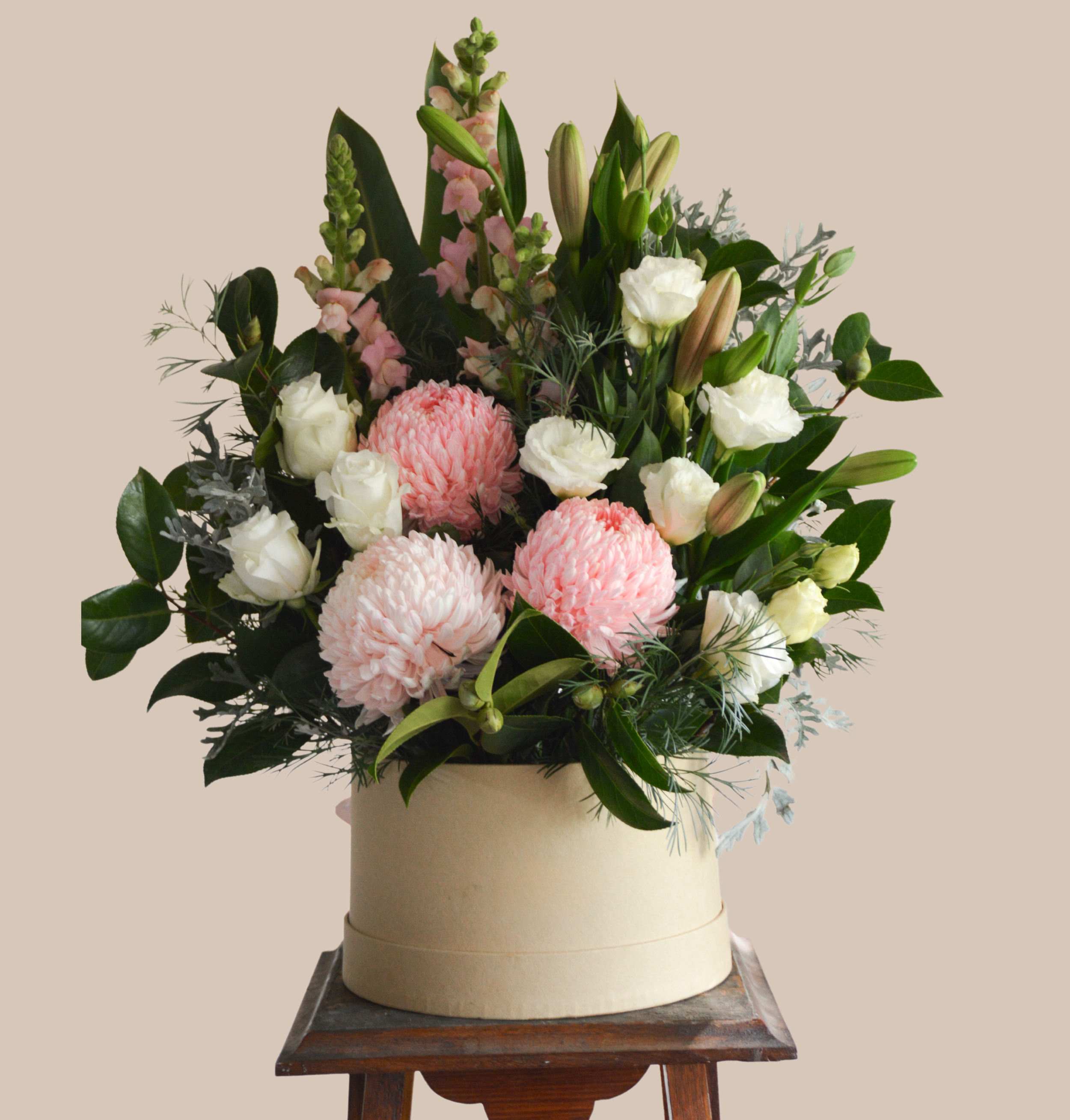 A floral arrangement with pink and white flowers in a round cream-colored hatbox on a wooden stand.