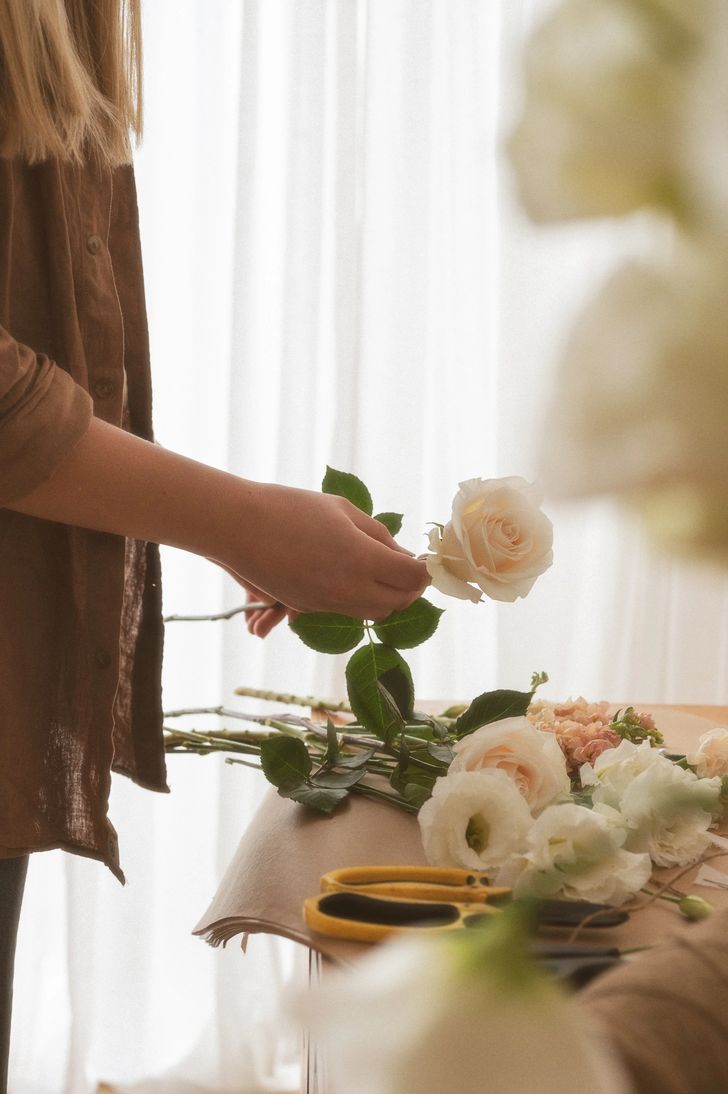 Person arranging white roses in a flower arrangement on a table with scissors and other flowers.
