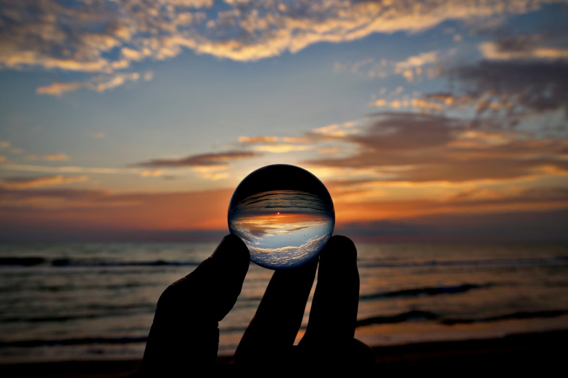 Person holding a glass sphere at sunset on the beach, with clouds and the sky reflected upside down in the sphere
