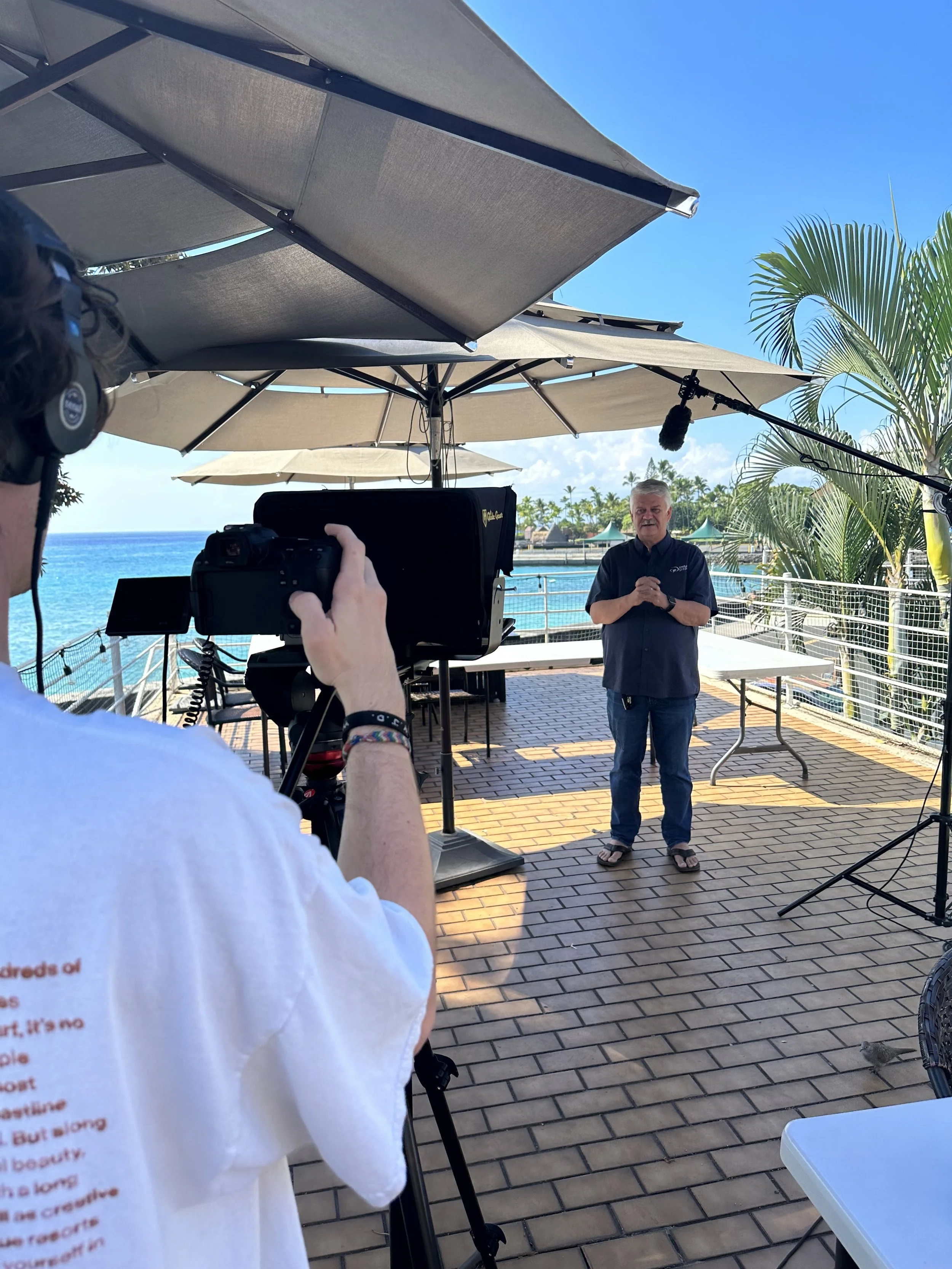 A man in a dark blue shirt and jeans standing on a rooftop patio, giving a speech or interview, with a large professional camera and crew recording him. The background shows a sunny day, blue sky, ocean, palm trees, and umbrellas.