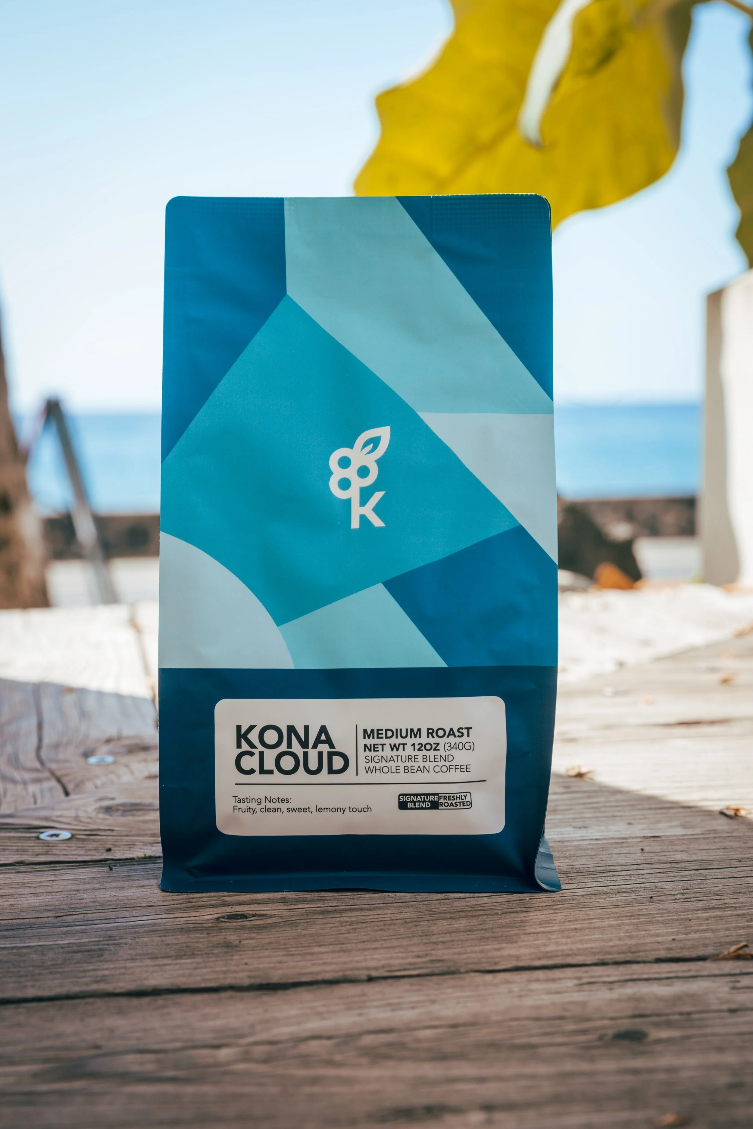 A bag of Kona Cloud medium roast whole bean coffee on a wooden table outdoors, with ocean and yellow leaves in the background.