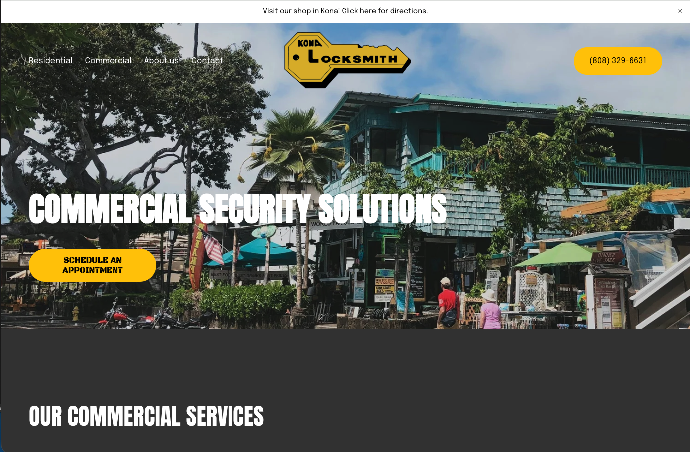 A commercial building with a blue-green exterior, surrounded by trees, with a sign for Kona Locksmith and a sticky note with a phone number. There are people walking by and outdoor tables with umbrellas in front of shops.