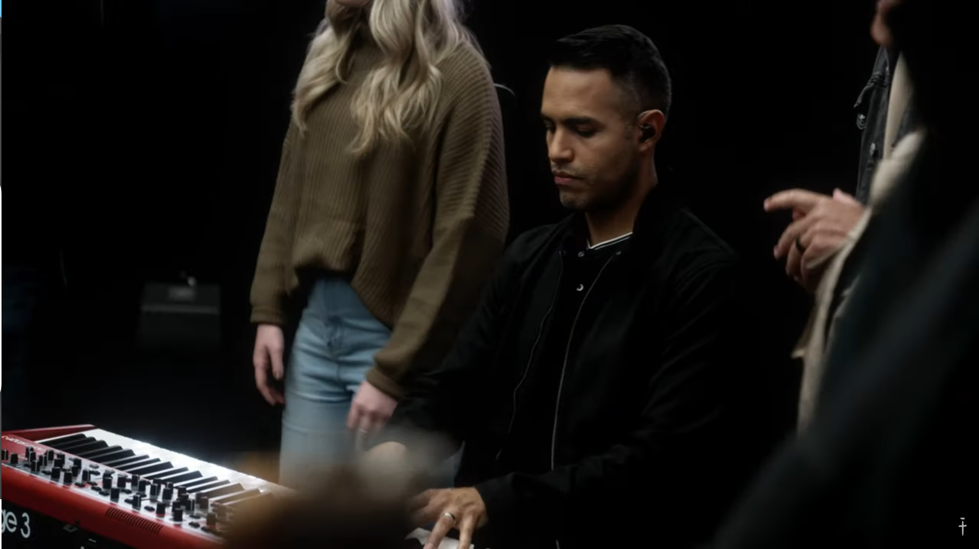 Man sitting at a keyboard with eyes closed, surrounded by people in a dark room.