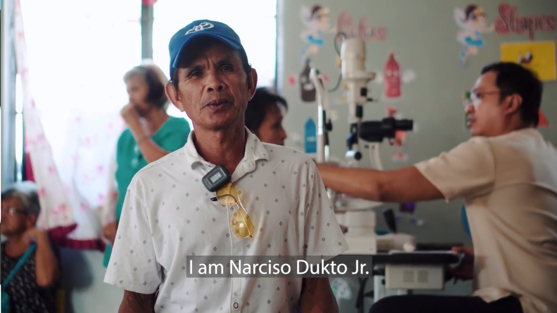 A middle-aged man wearing a blue cap and glasses hanging around his neck introduces himself as Narciso Dukto Jr. in a hospital room with medical staff and patients in the background.