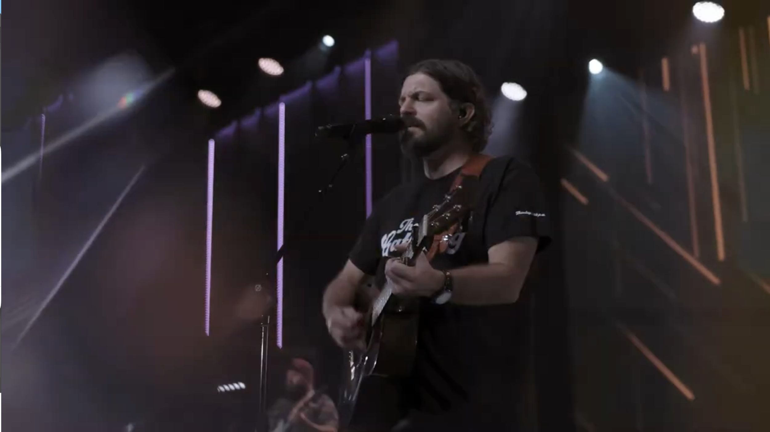 A man singing into a microphone while playing an acoustic guitar on stage with colorful stage lights in the background.