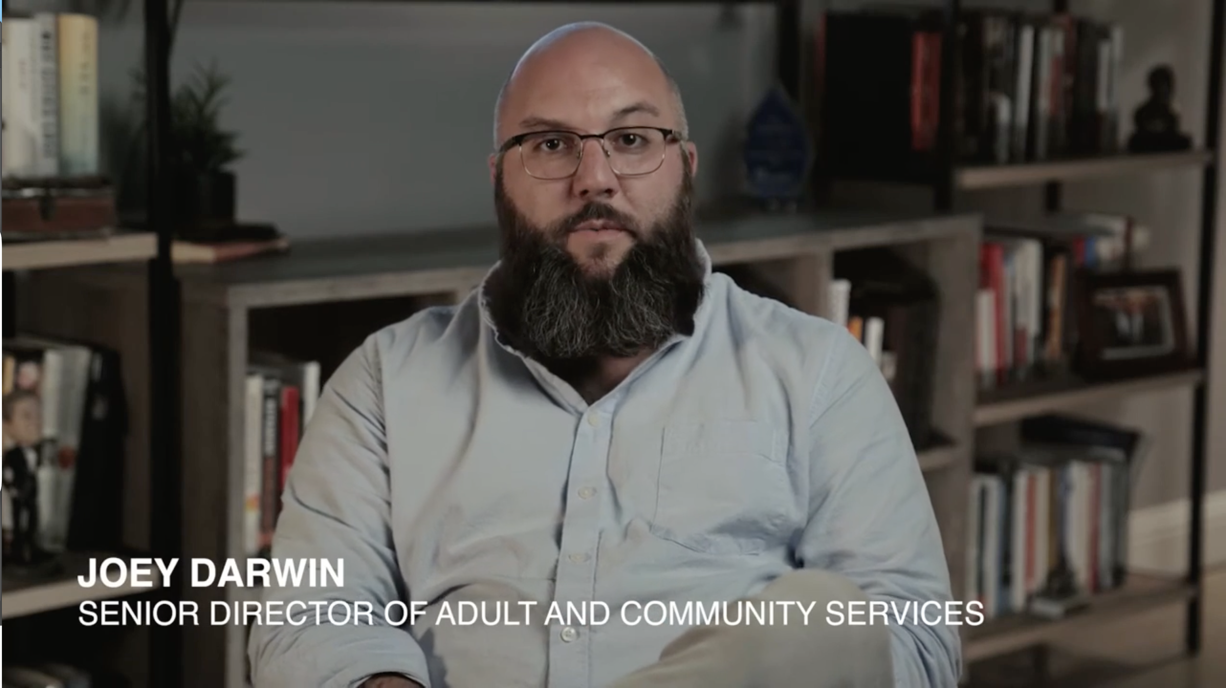 A man with glasses, a shaved head, and a full beard, wearing a light-colored shirt, sitting in front of a bookshelf filled with books.