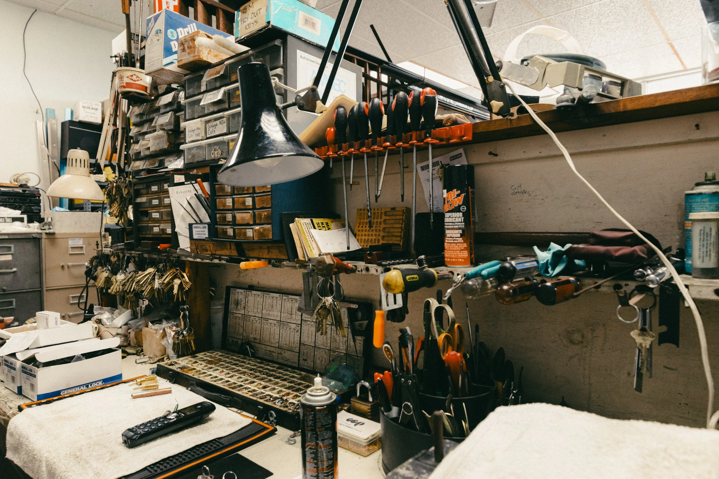 A cluttered workbench with various tools, keys, scissors, and supplies for repairs and projects, including screwdrivers, pliers, a remote, and small parts organized in containers and on hooks.