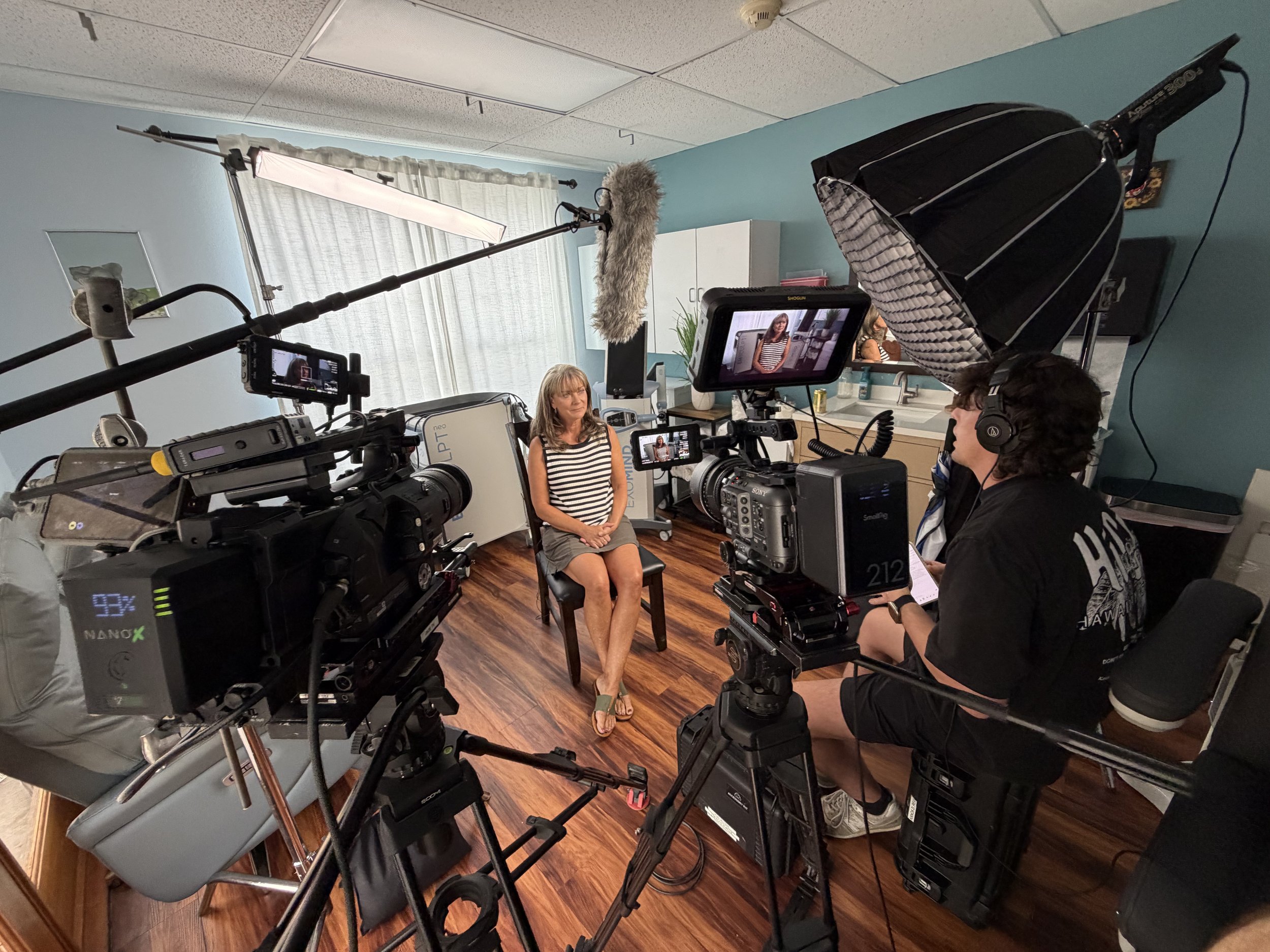 A woman sitting on a chair during a video shoot or interview, surrounded by cameras, lighting equipment, and crew members in a small room with blue walls and hardwood floors.