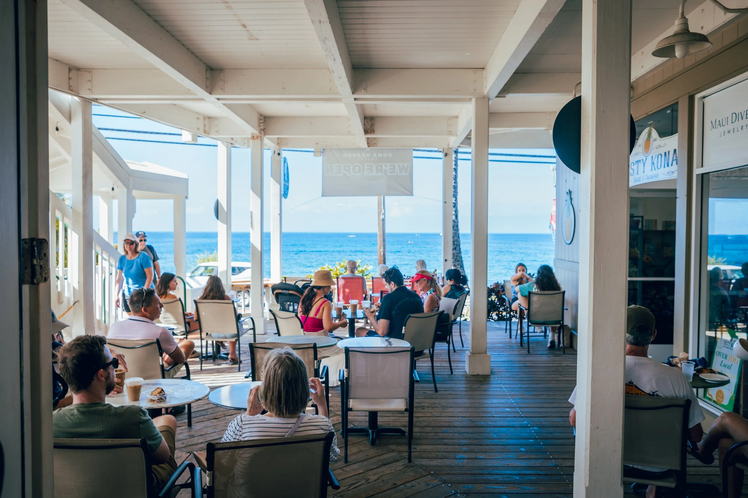 People dining on a beachside patio overlooking the ocean, with some seated at tables and others walking by, under a wooden structure with a view of the sea in the background.