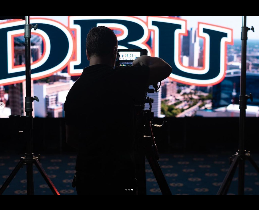 Photographer taking pictures of a cityscape on a large illuminated sign that reads 'OBU' with studio lights visible.