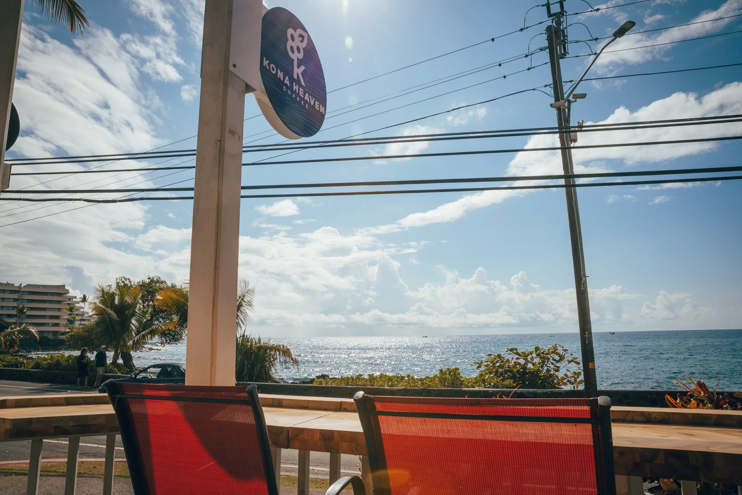 Seaside view from a cafe with red chairs, palm trees, and a large sign that reads 'Kona Heaven Coffee,' overlooking the ocean, with power lines and a blue sky with clouds.
