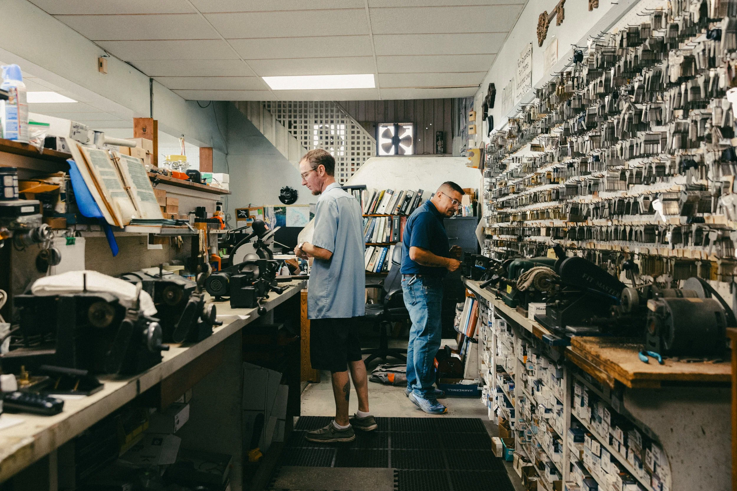 Two men working in a repair shop or hardware store, surrounded by tools, equipment, and organized small parts and hardware on the walls and shelves.