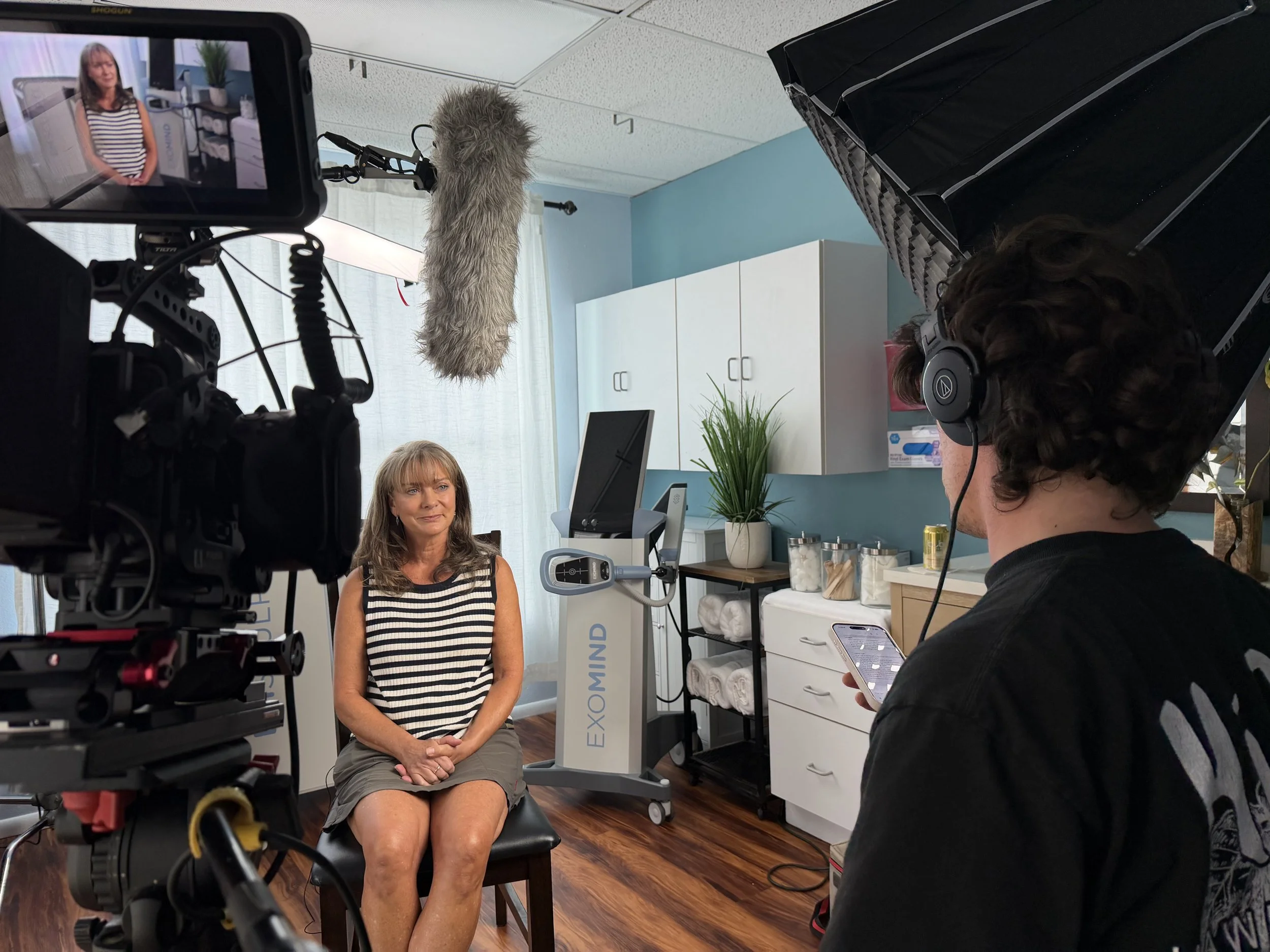 A woman in a striped sleeveless top and gray skirt is being filmed in a studio or medical setting with a camera and crew. Behind her is a blue wall, white cabinets, a large potted plant, and a medical device labeled brand 'EXOMIND'. The crew member, 