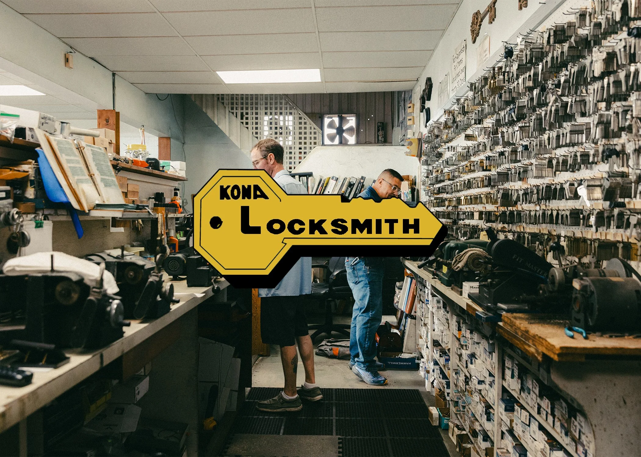 The interior of a locksmith shop with two men working among shelves filled with lock keys, tools, and equipment. There's a yellow sign with black text that reads 'Kona Locksmith' in the foreground.