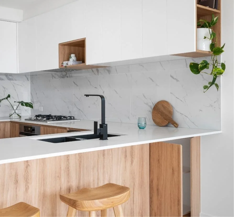 Modern kitchen with white and wood cabinets, marble backsplash, black faucet, small glass, and wooden cutting board on white countertop, with green plants.