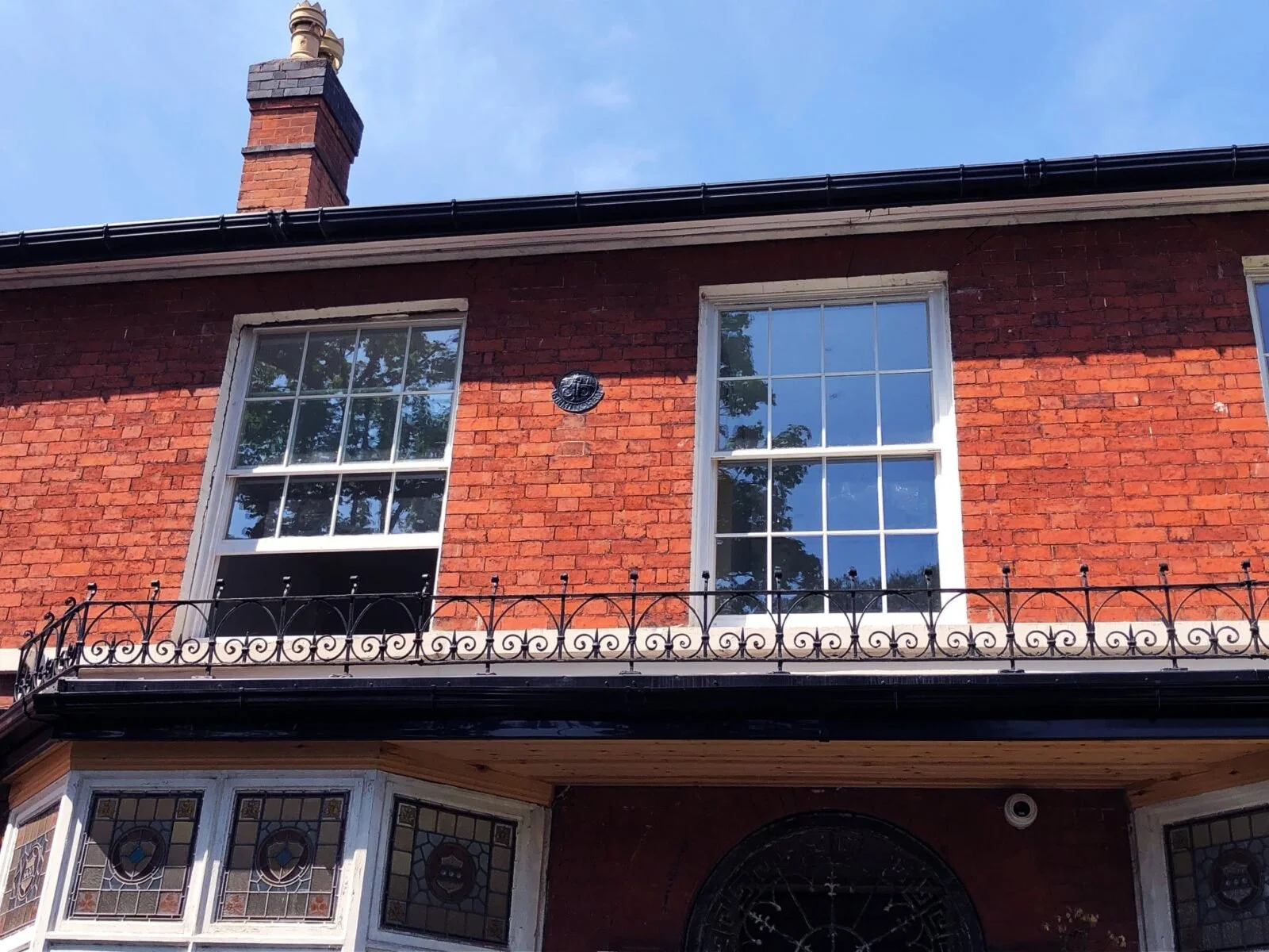A brick house with two large white-framed windows on the second floor, a decorative wrought iron balcony railing, and a Victorian-style bay window with stained glass on the first floor. There is a chimney on the roof and a black gutter along the edge of the roof. The sky is clear and blue.