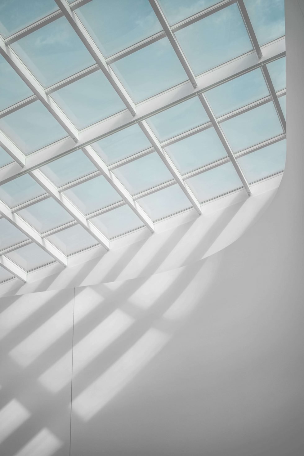 Interior view of a modern building with large glass roof panels creating geometric shadows on a white wall.
